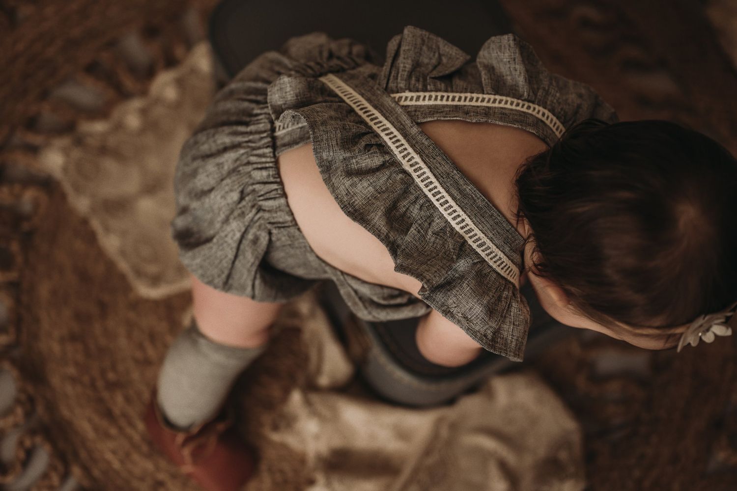 A vintage-themed photo of a toddler in a gray dress sitting on textured fabric from above.
