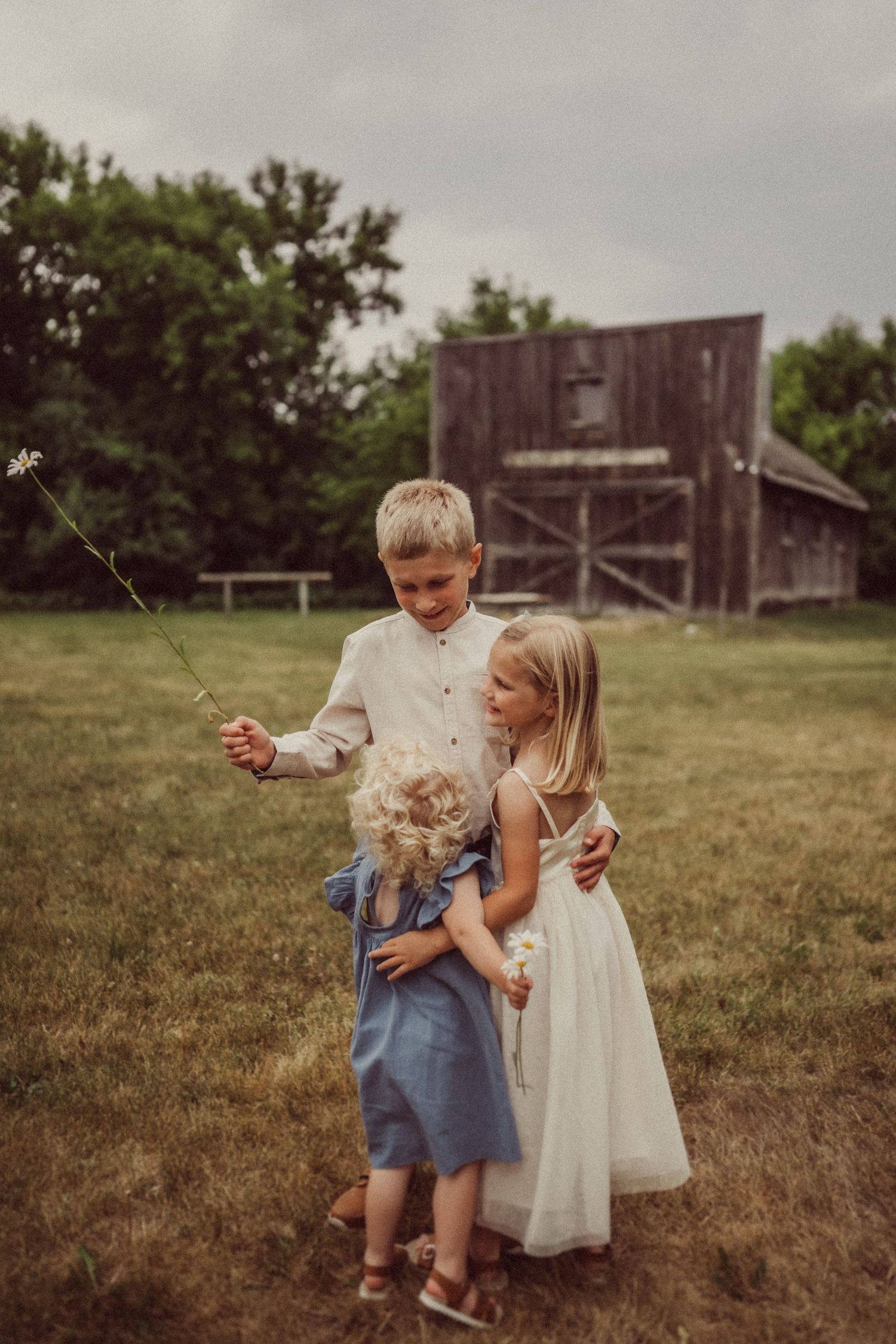 Children stand together in a field holding flowers with a rustic barn in the background.