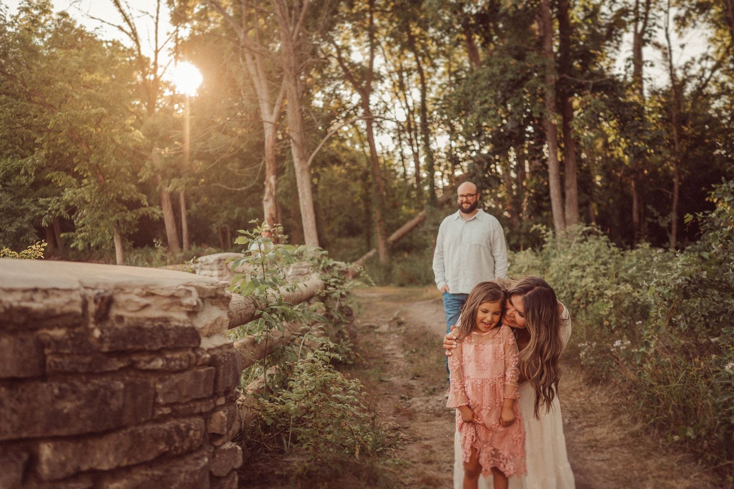Couple walking along stone wall path in wooded park during sunset.
