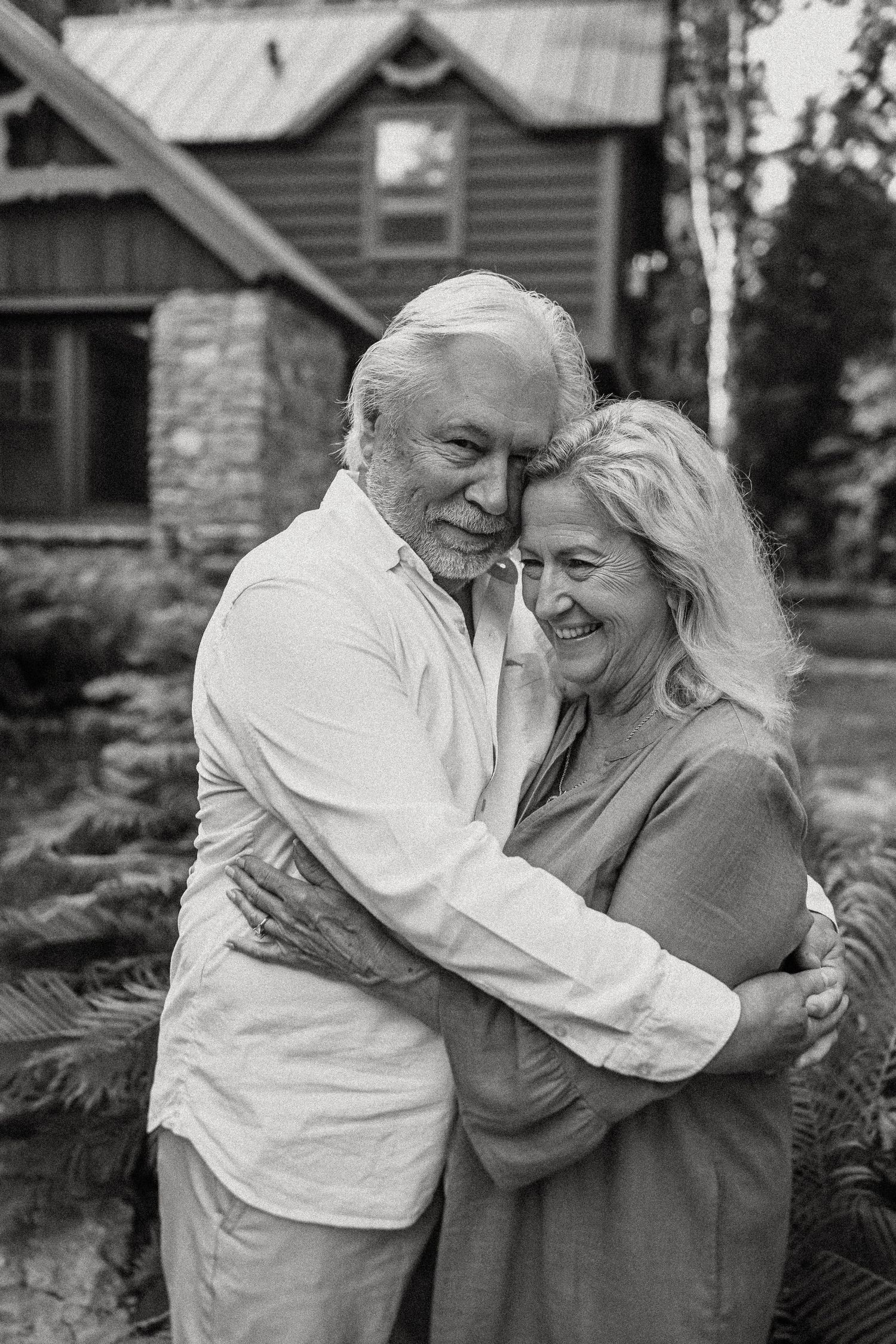 Loving elderly couple embrace while laughing together in front of rustic cabin with stone accents.