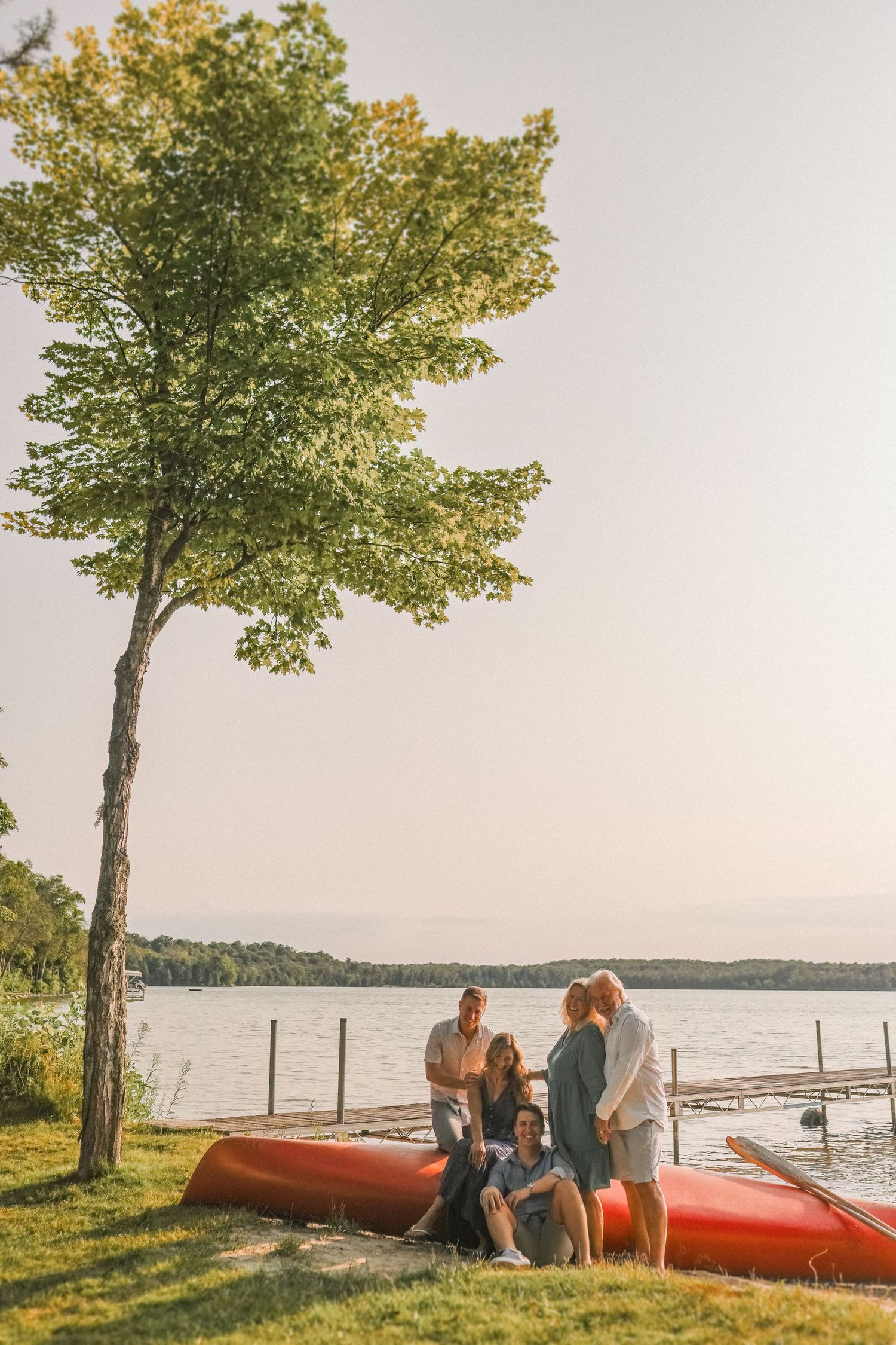 A family relaxes by red kayaks near a lakeside tree during golden hour.