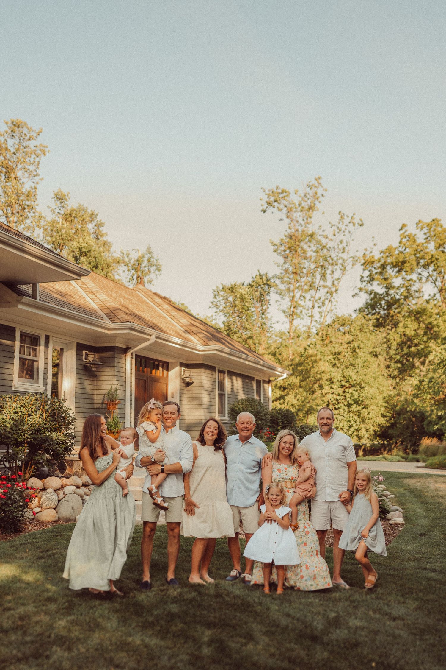 Extended family posing for a warm sunset photo in coordinated white and beige outfits by their home.