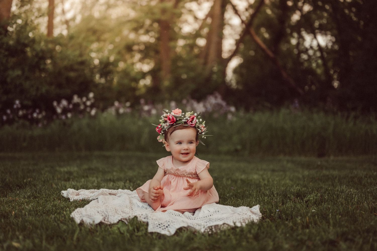 A small child wearing a floral crown sits on a white blanket in a sunlit garden.