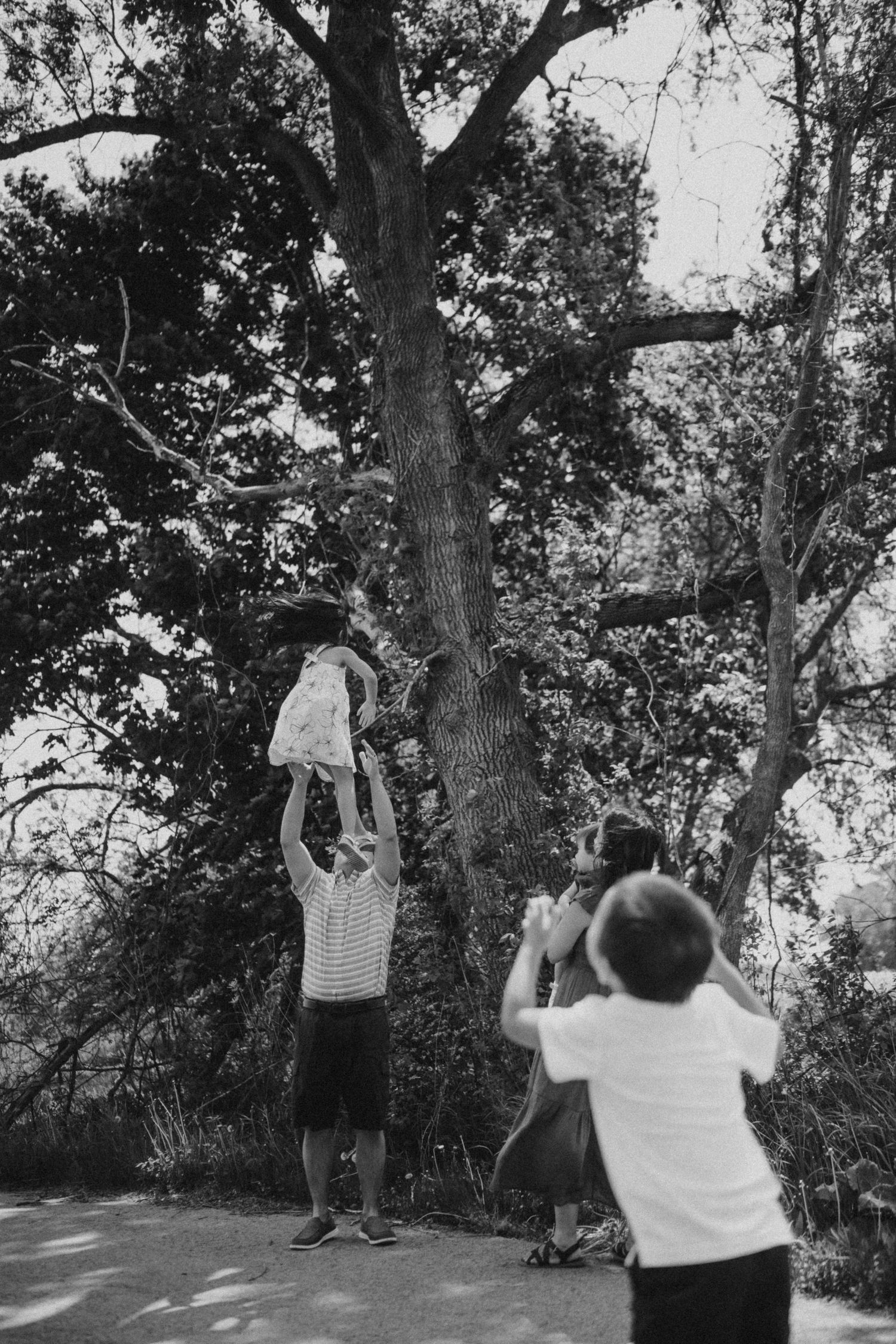 A group plays together under a tall tree, with someone lifting a child holding a basket.