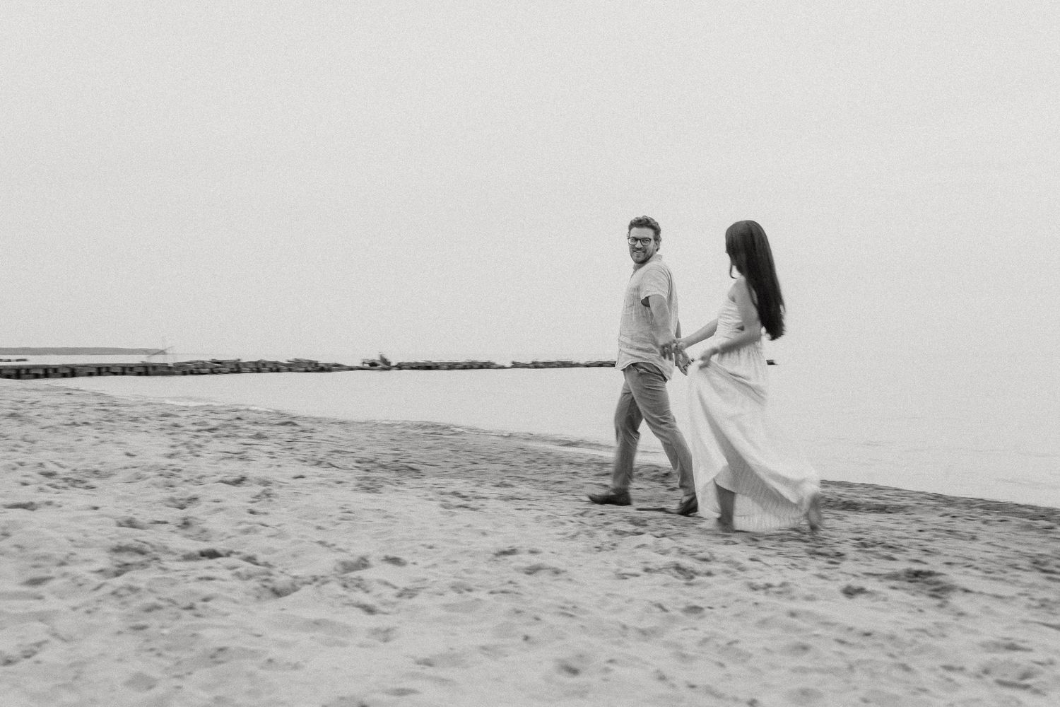 Black and white photo of couple strolling along misty beach shoreline.