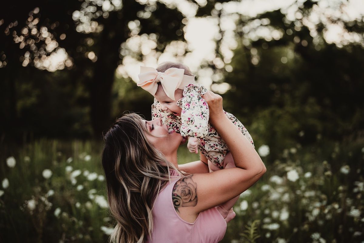 Woman in pink dress lifting baby in floral outfit among blooming wildflowers.