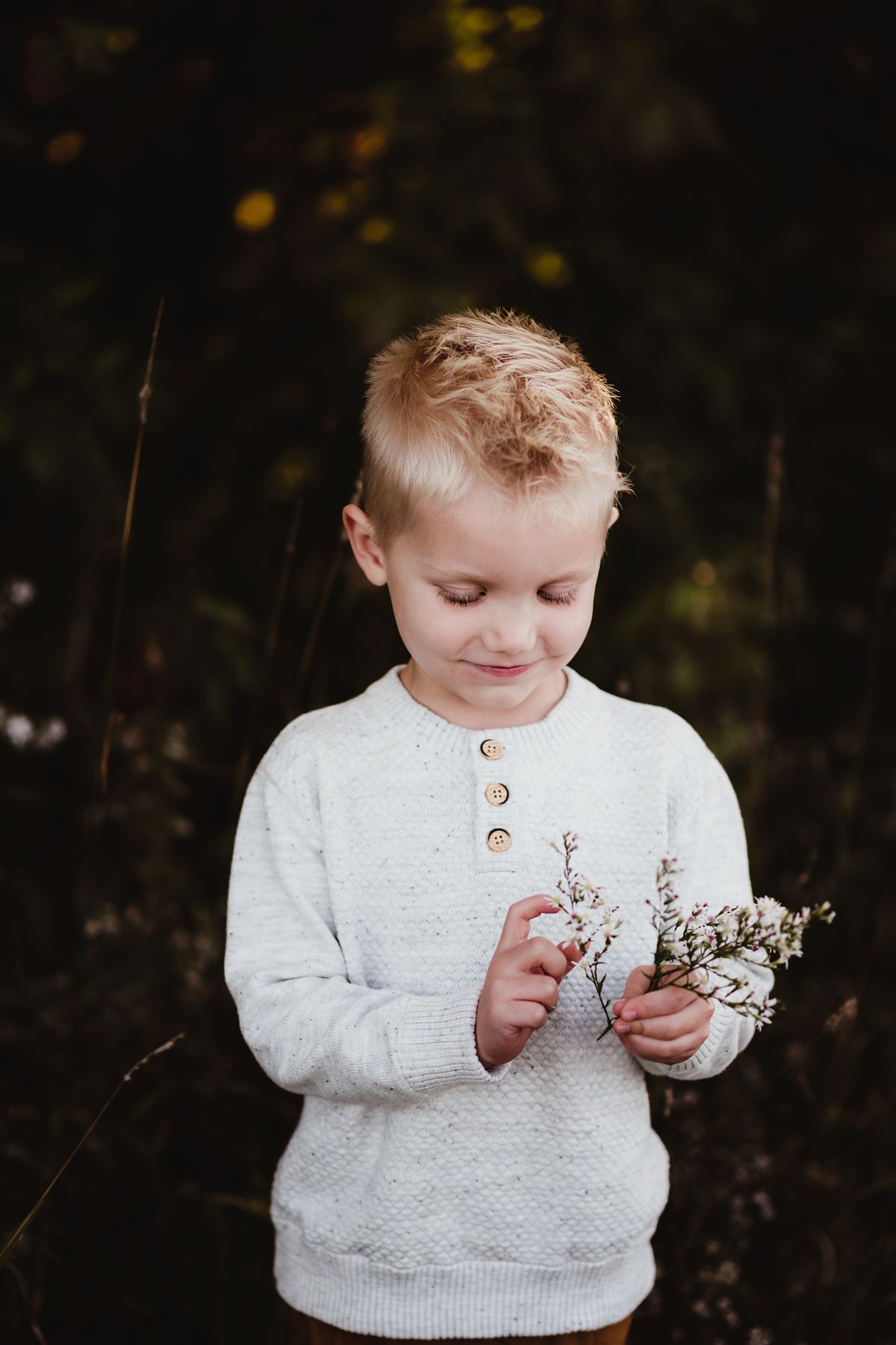 A young child in a white sweater gently examines small white flowers outdoors.
