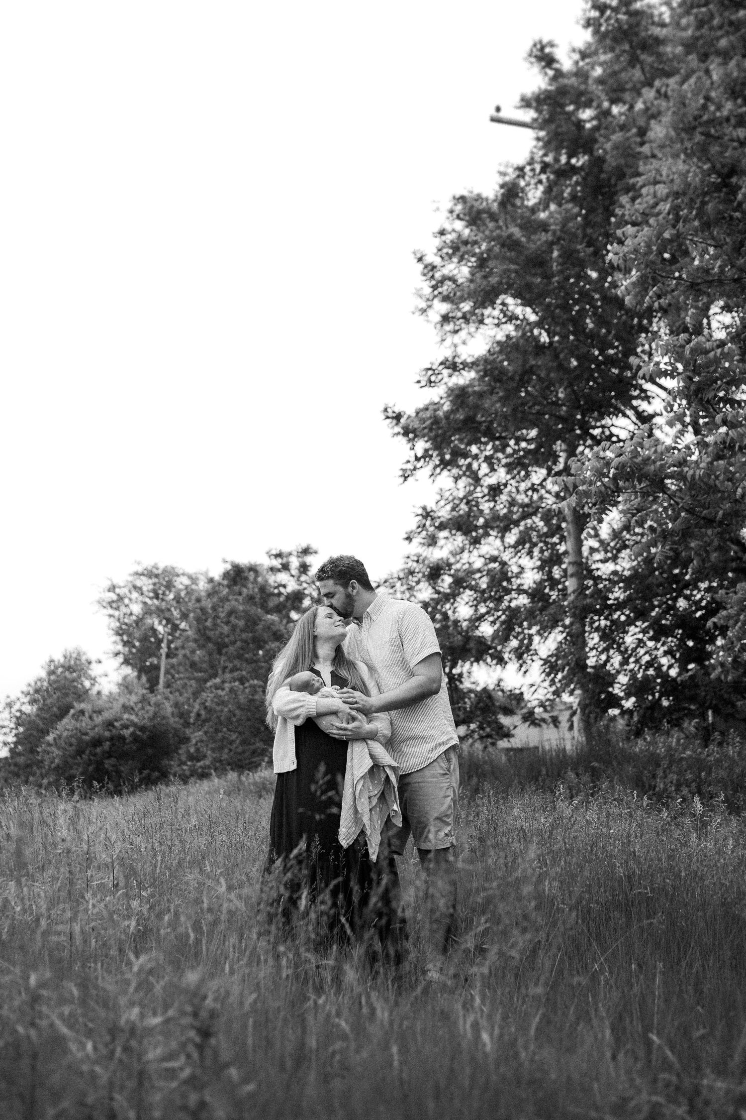 Black and white image of two people embracing in a grassy field beneath tall trees.