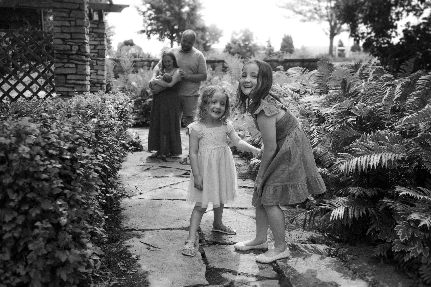 Vintage black and white photograph showing people dancing together on a garden path surrounded by lush plants and foliage.