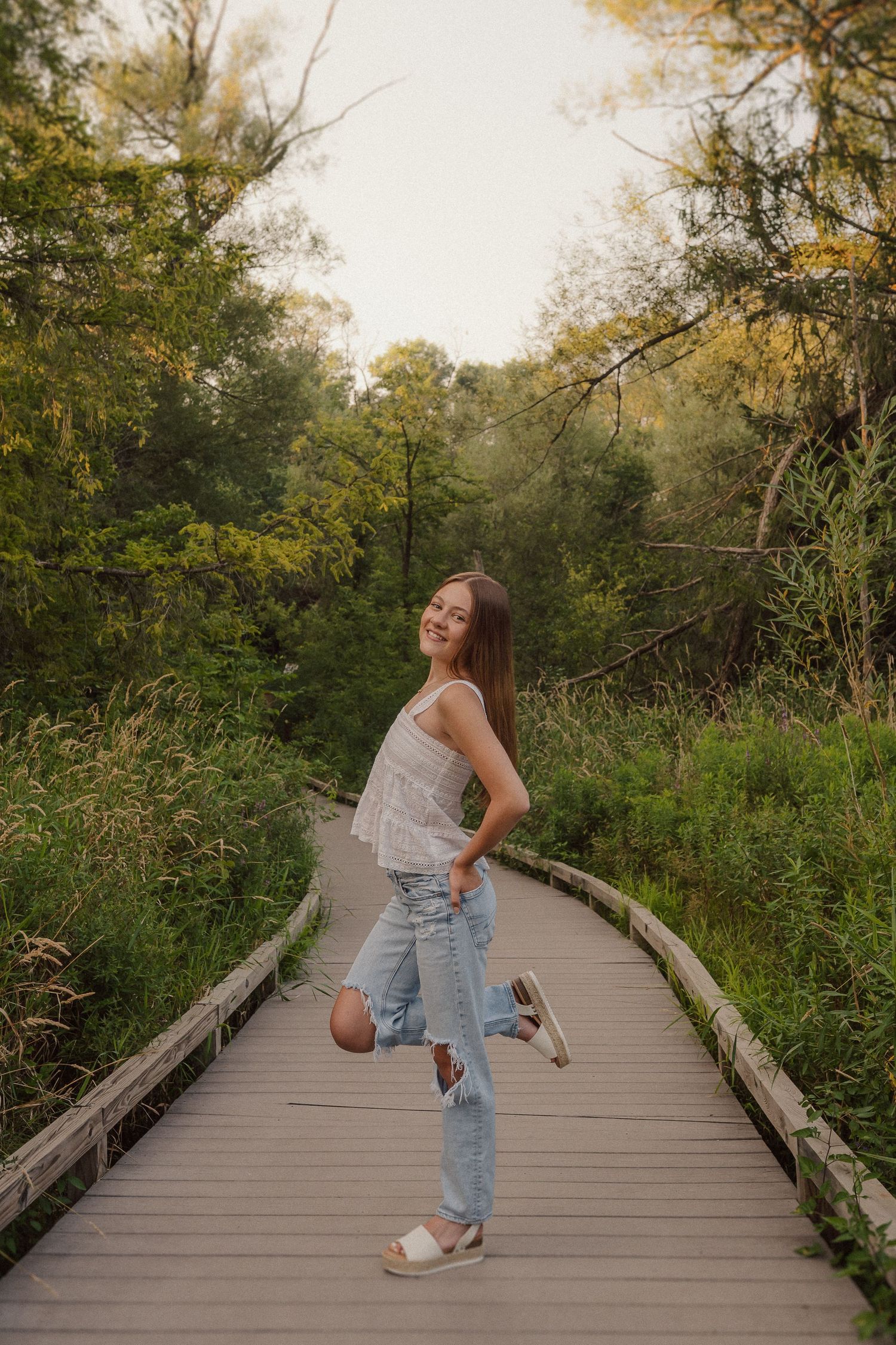 A person wearing jeans and a white top poses casually on a wooden boardwalk surrounded by greenery.