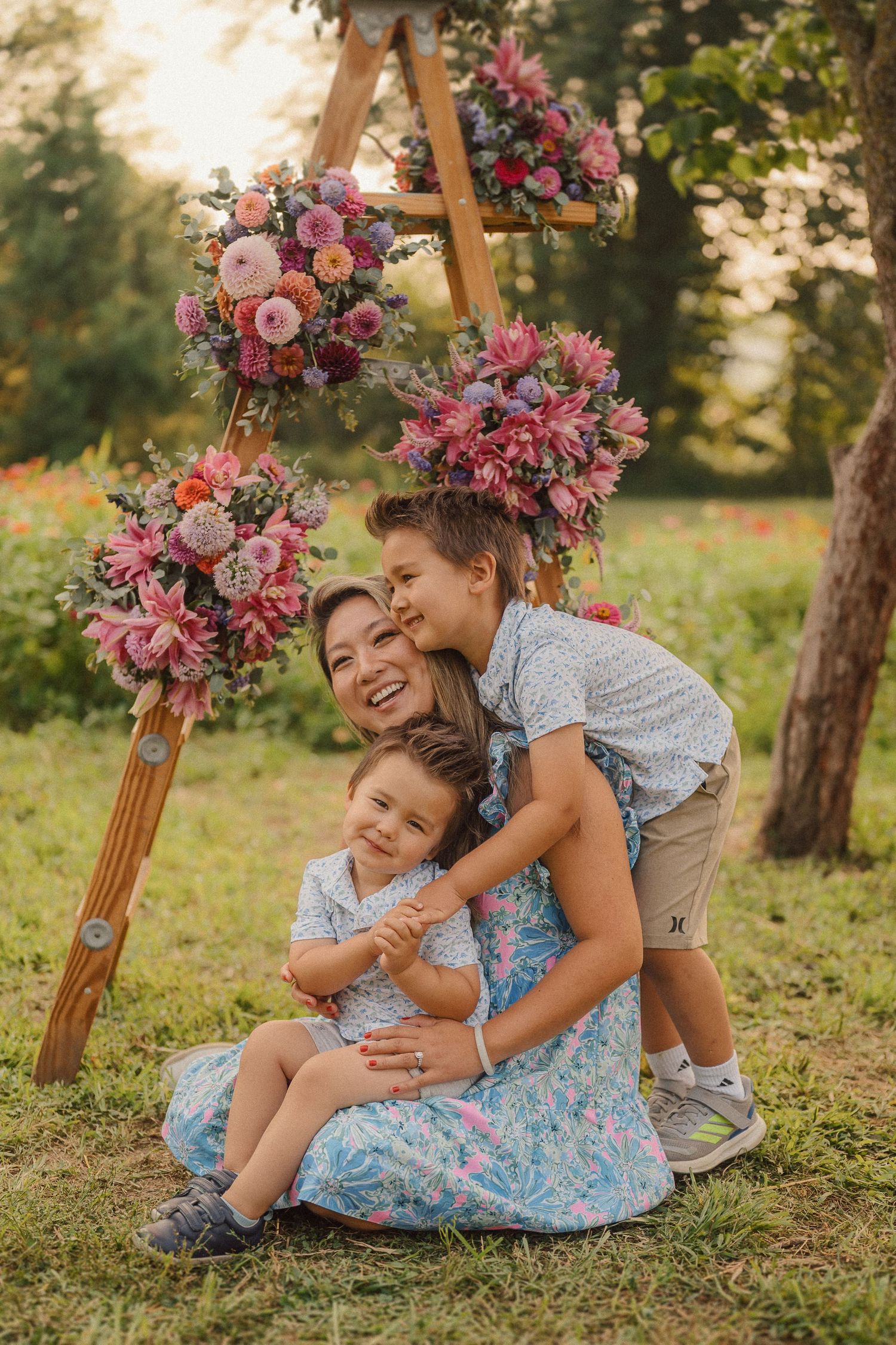 A sweet family moment captured under a wooden ladder decorated with pink and purple flower arrangements outdoors.