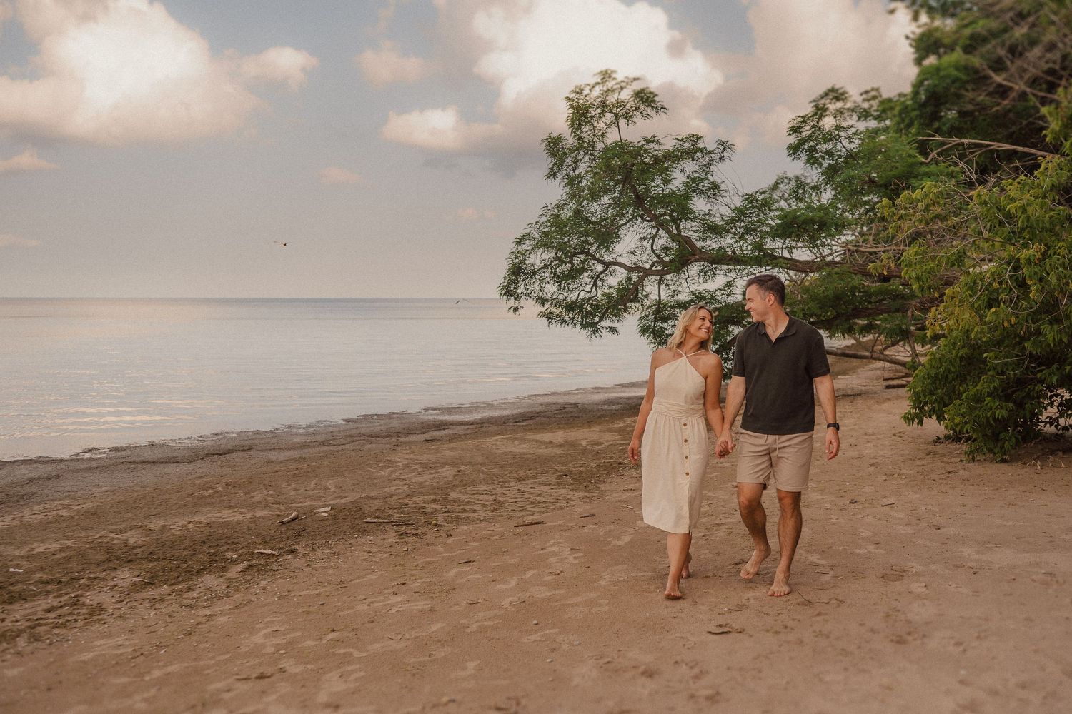 A couple strolls hand in hand along a tropical beach at sunset, surrounded by lush trees and sandy shoreline.