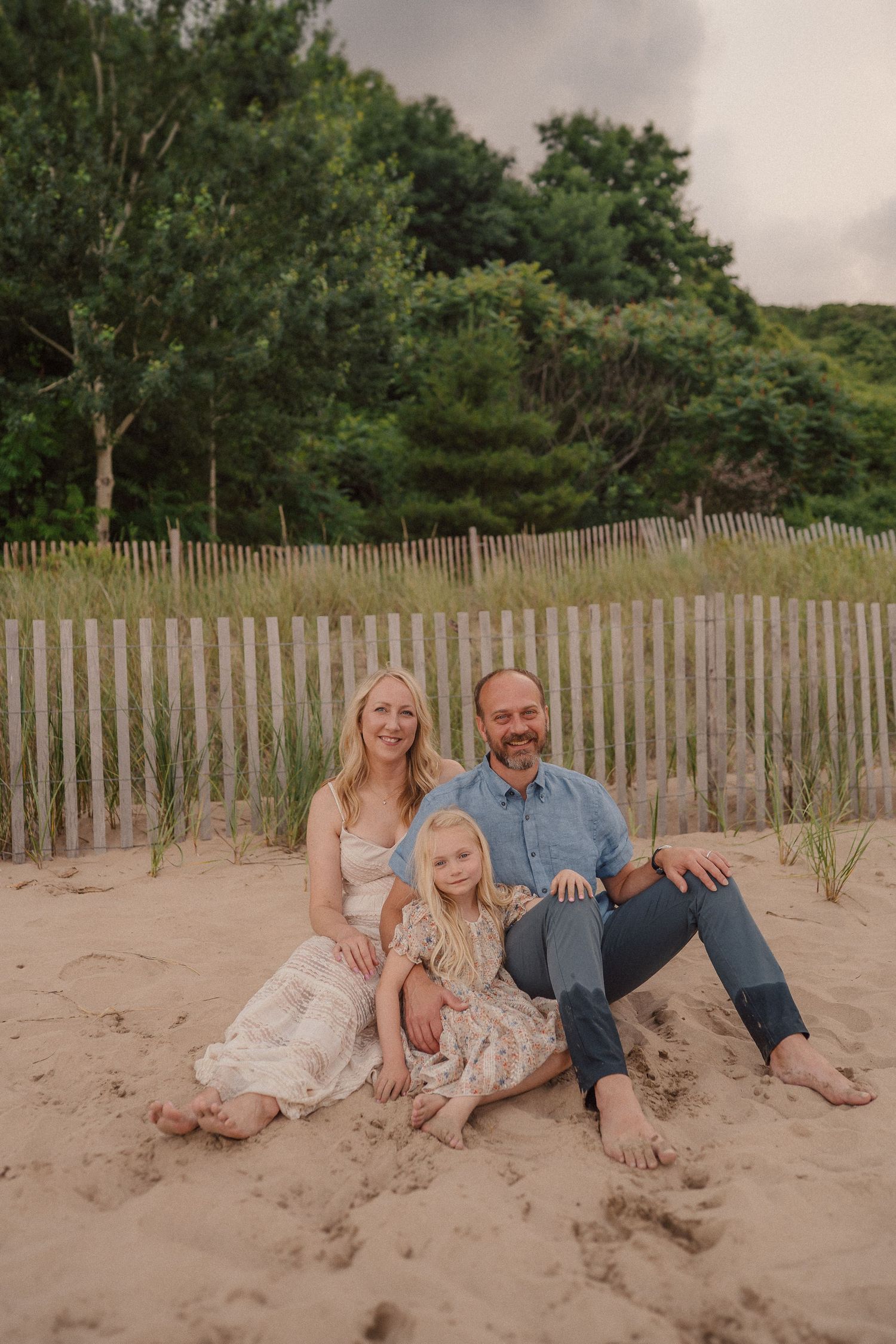 A family sits together on a sandy beach with weathered wood fencing and green foliage in the background at sunset.