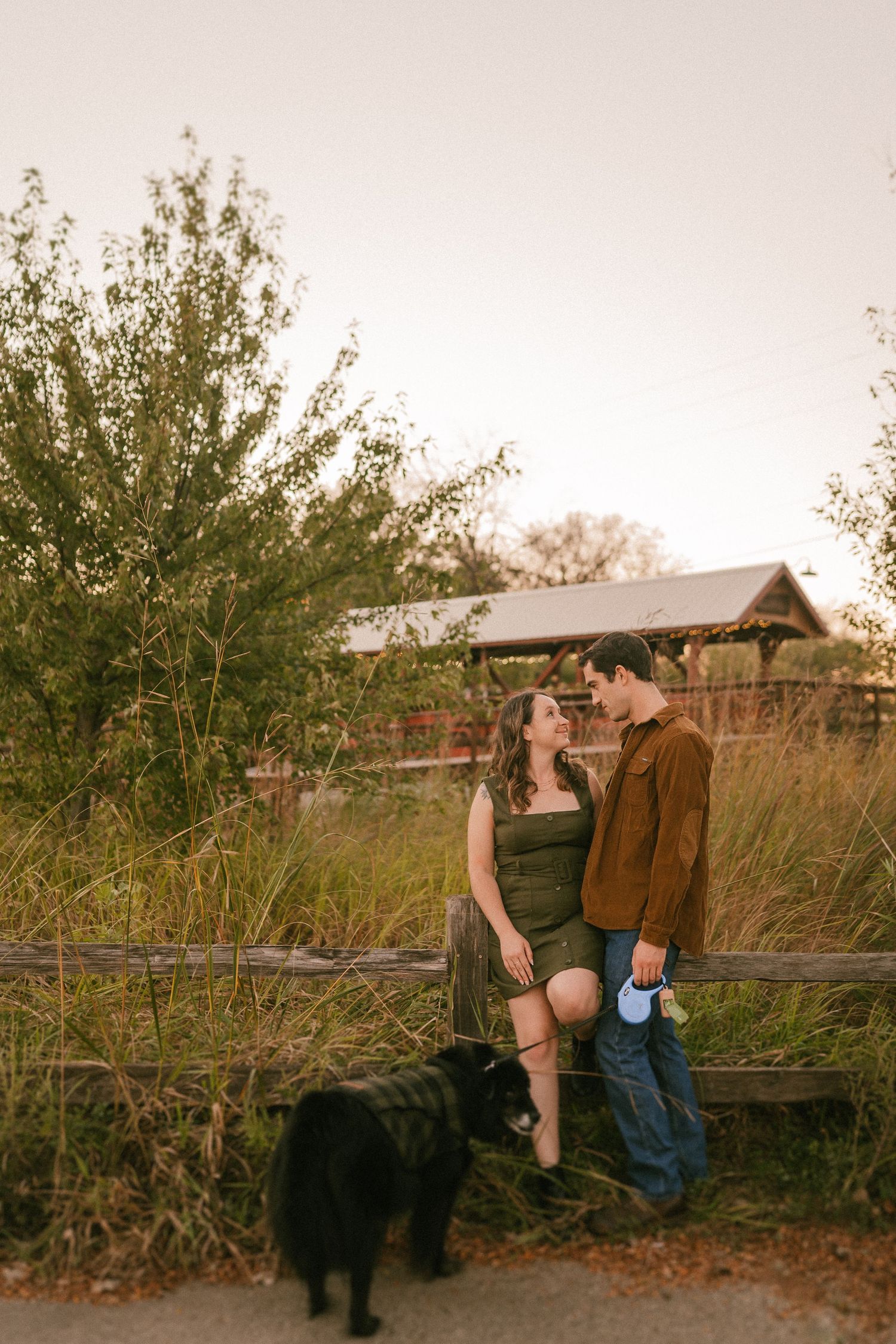 A couple and their black dog pause for a moment by a wooden fence during sunset.