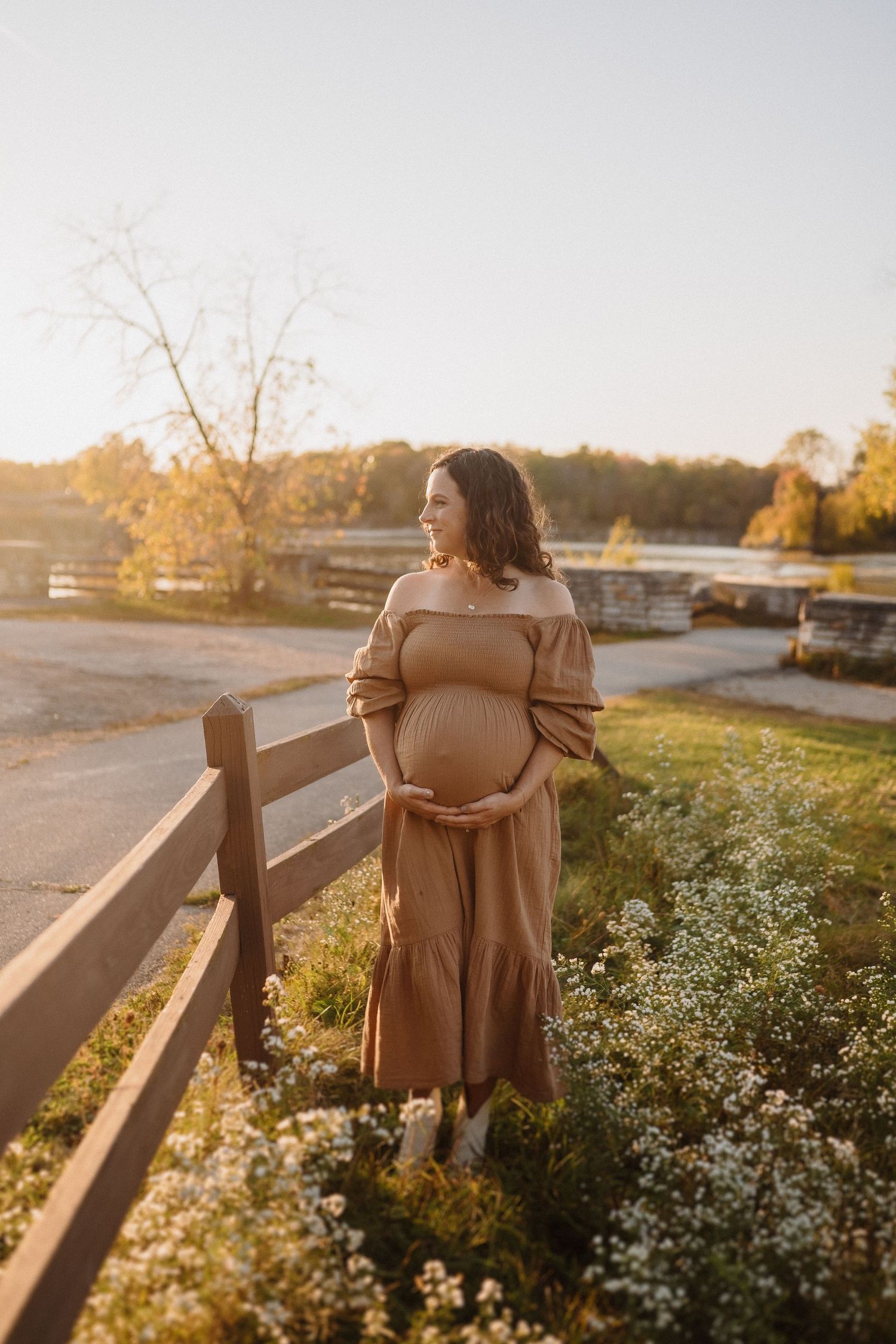 Pregnant woman in brown dress gazes across landscape by wooden fence at sunset.