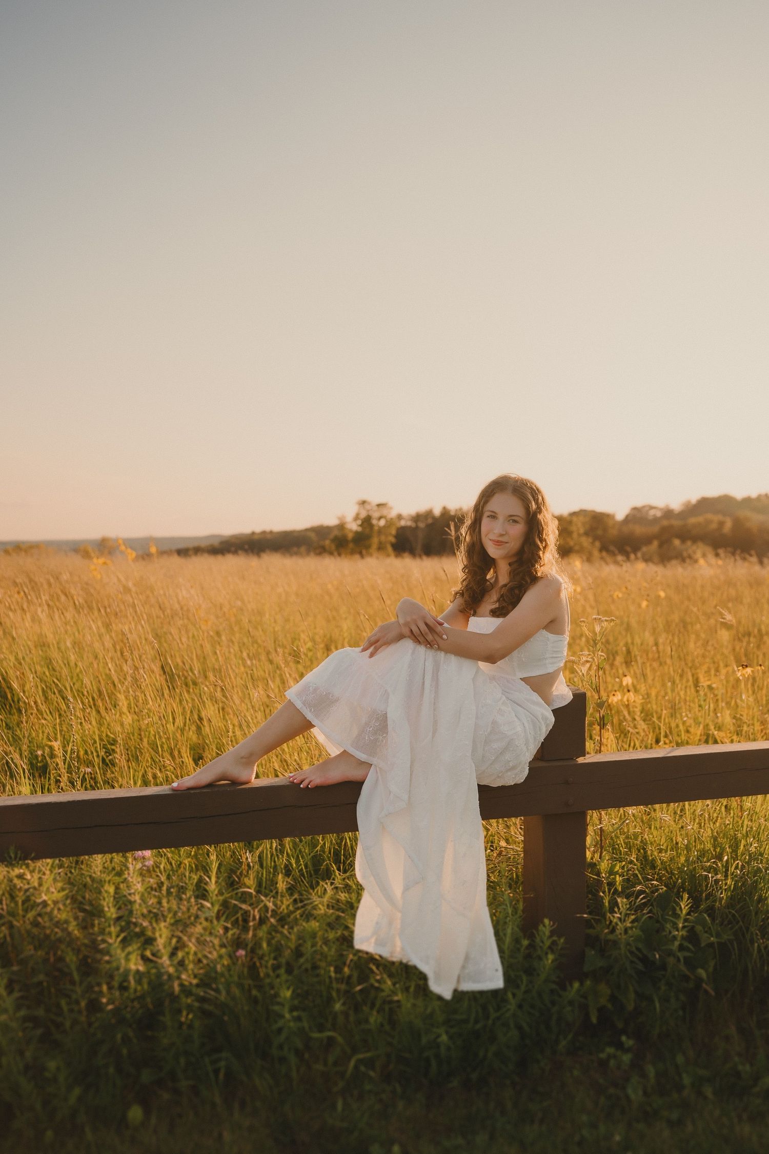 A person in a white dress sits on a wooden fence at sunset in a golden field of tall grass.
