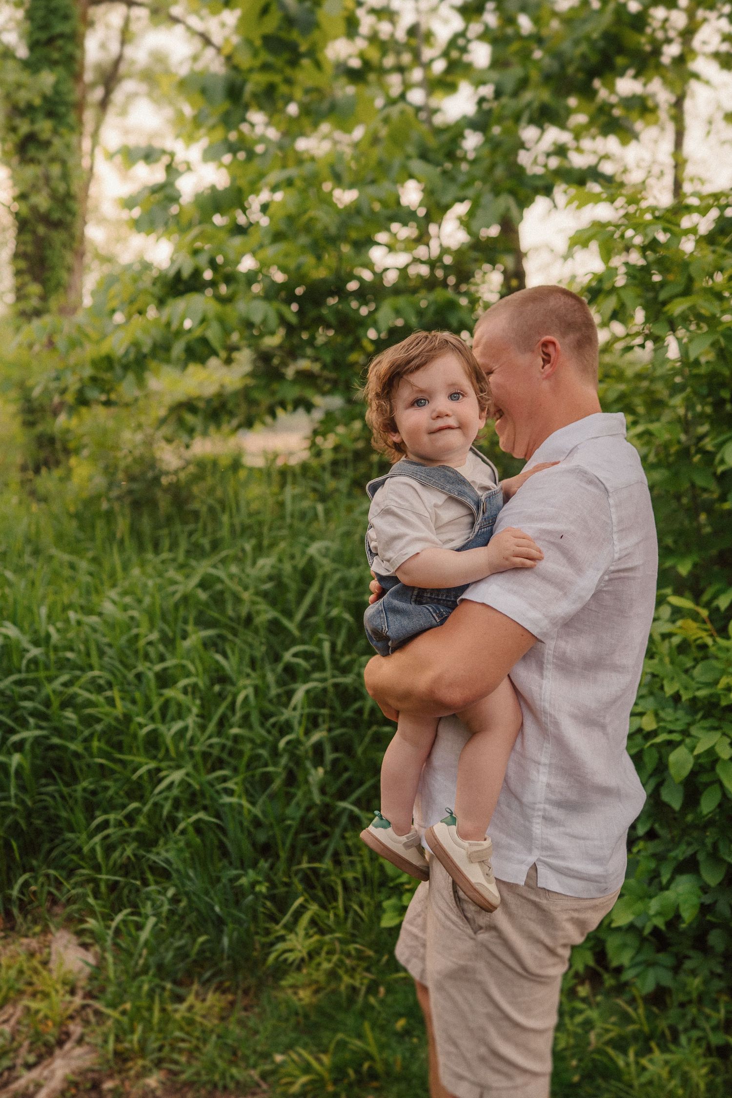 Parent holding child in casual summer attire against lush green foliage background.