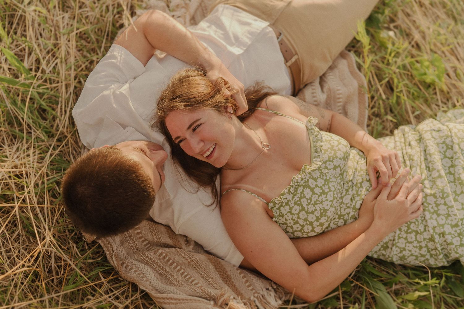 A romantic couple lies on a blanket in tall grass during a golden hour outdoor photoshoot.