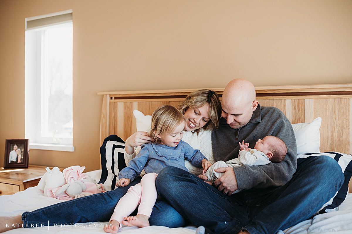 Family cuddle together on bed while reading to toddler and holding newborn.