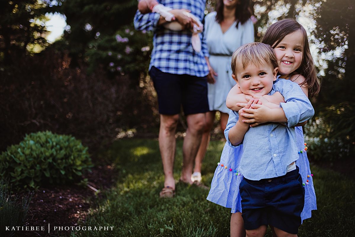 Two children in blue outfits embrace while playing outdoors in a garden setting at sunset.