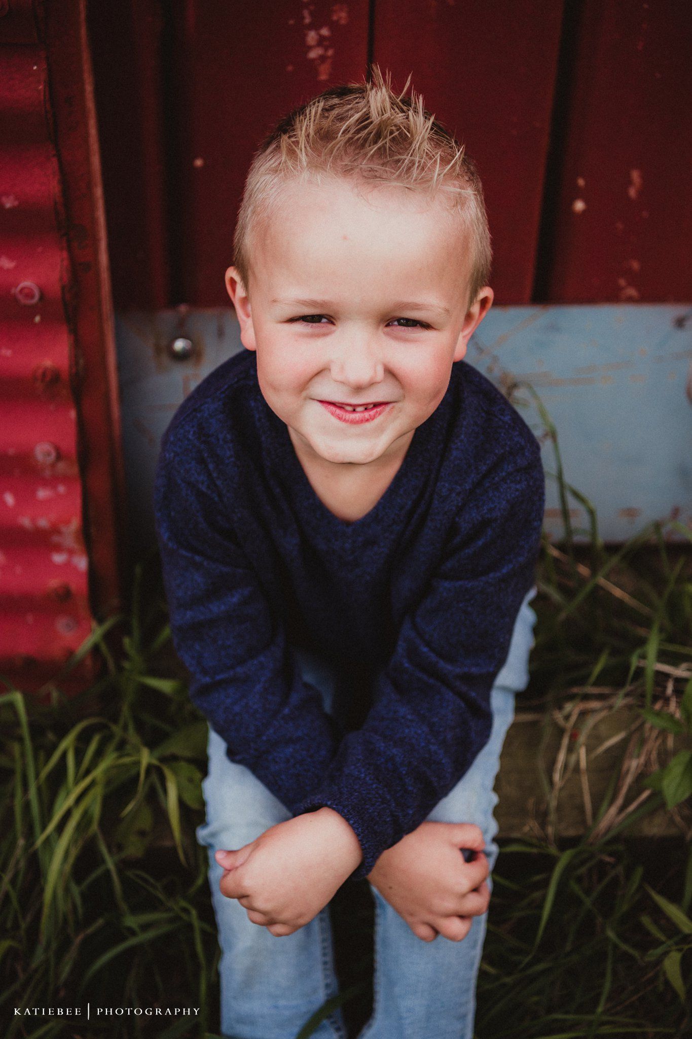 Candid portrait of a young child in a navy sweater smiling against a red barn wall background.
