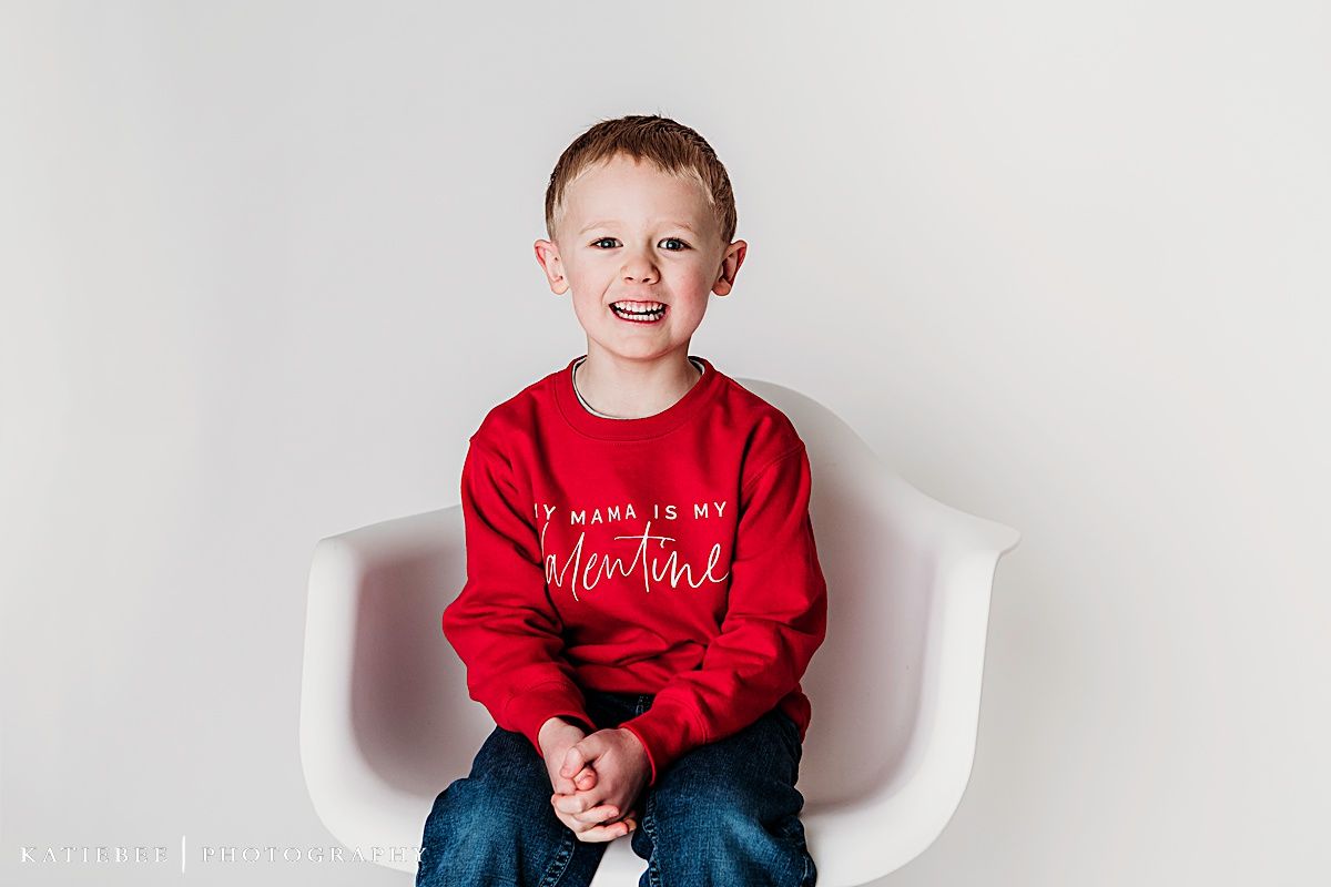 Cheerful young one wearing a red Valentine's sweater sitting on a modern white chair against light backdrop.