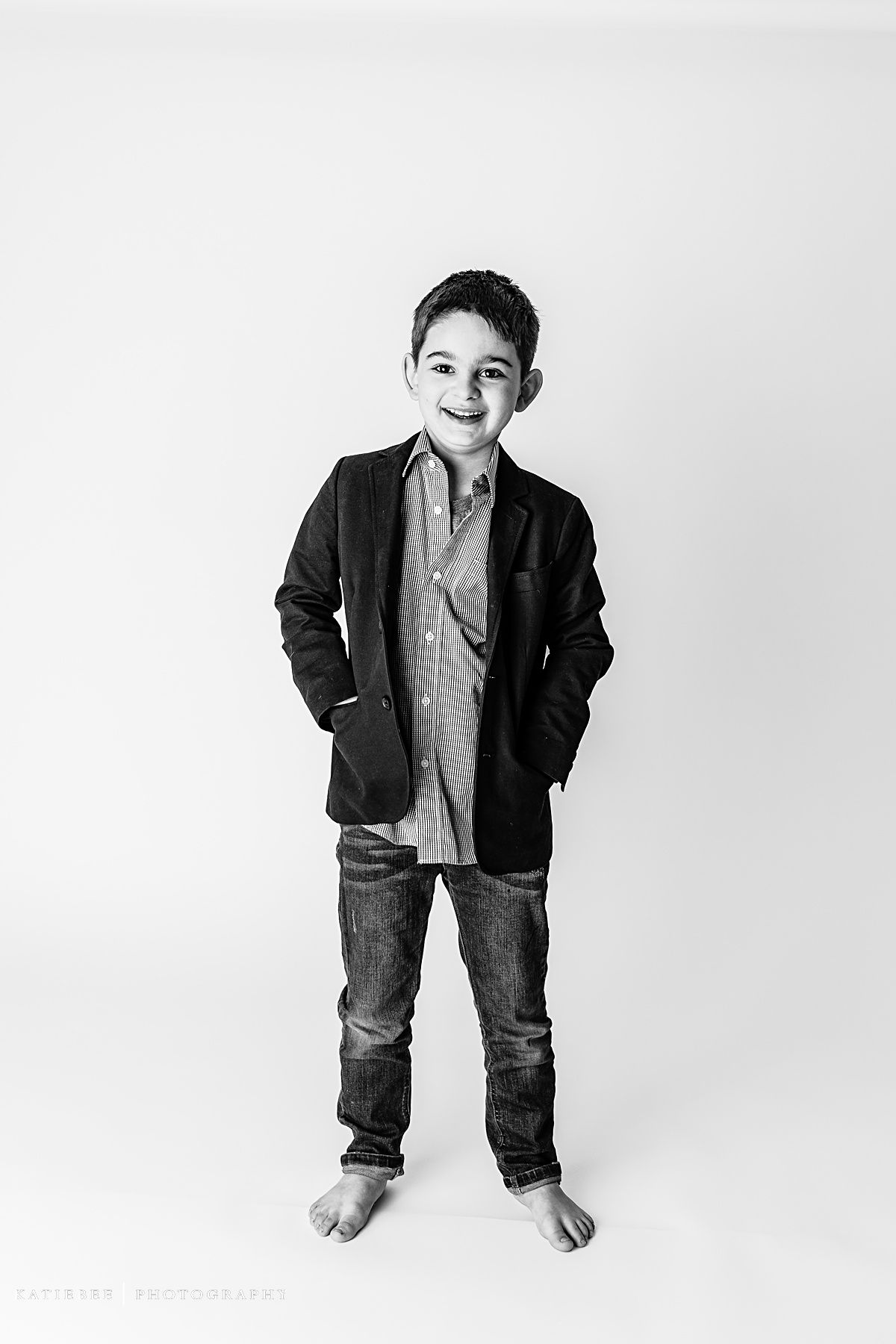 Black and white studio photo of a child in casual business attire standing barefoot against white background.