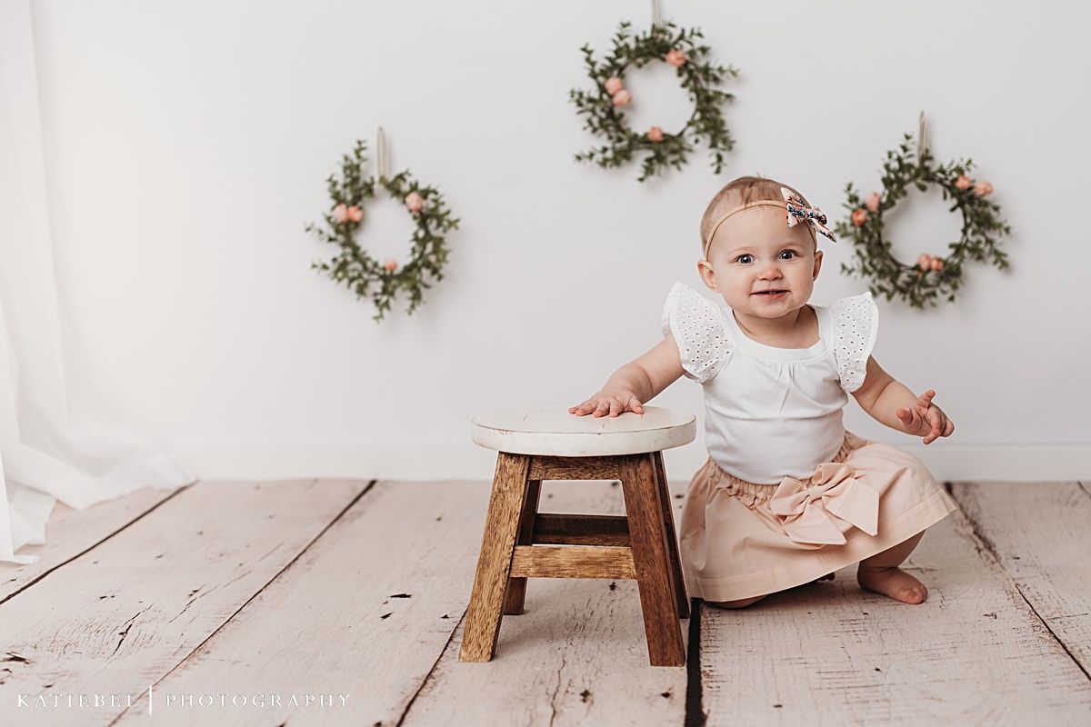Baby in white dress sitting near wooden stool against white background with wreaths.
