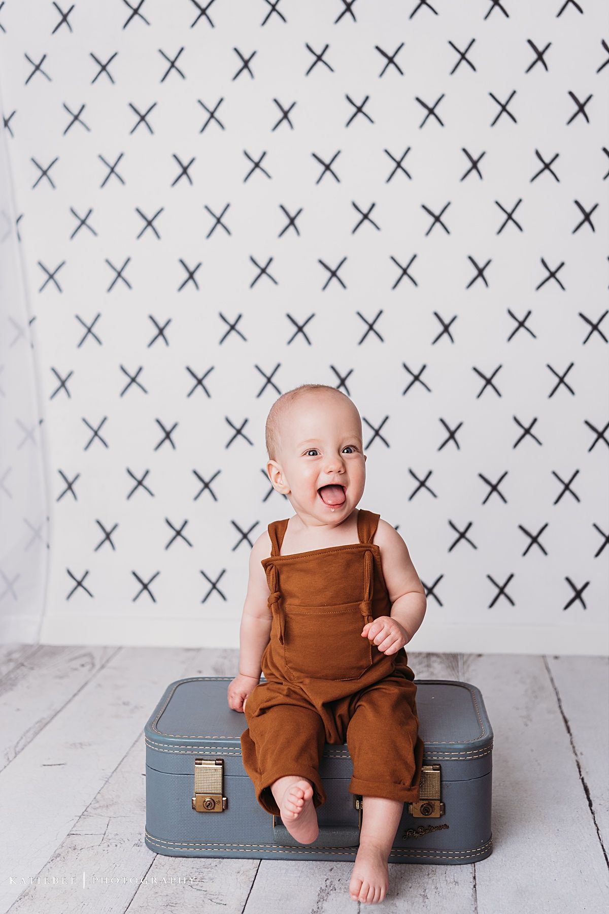 Laughing baby in brown overalls sits on vintage gray suitcase against X-pattern wallpaper background.