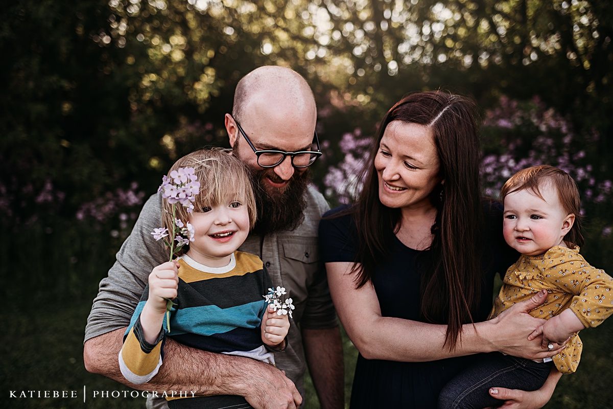 Joyful outdoor family portrait with purple flowers and natural sunset lighting in background.