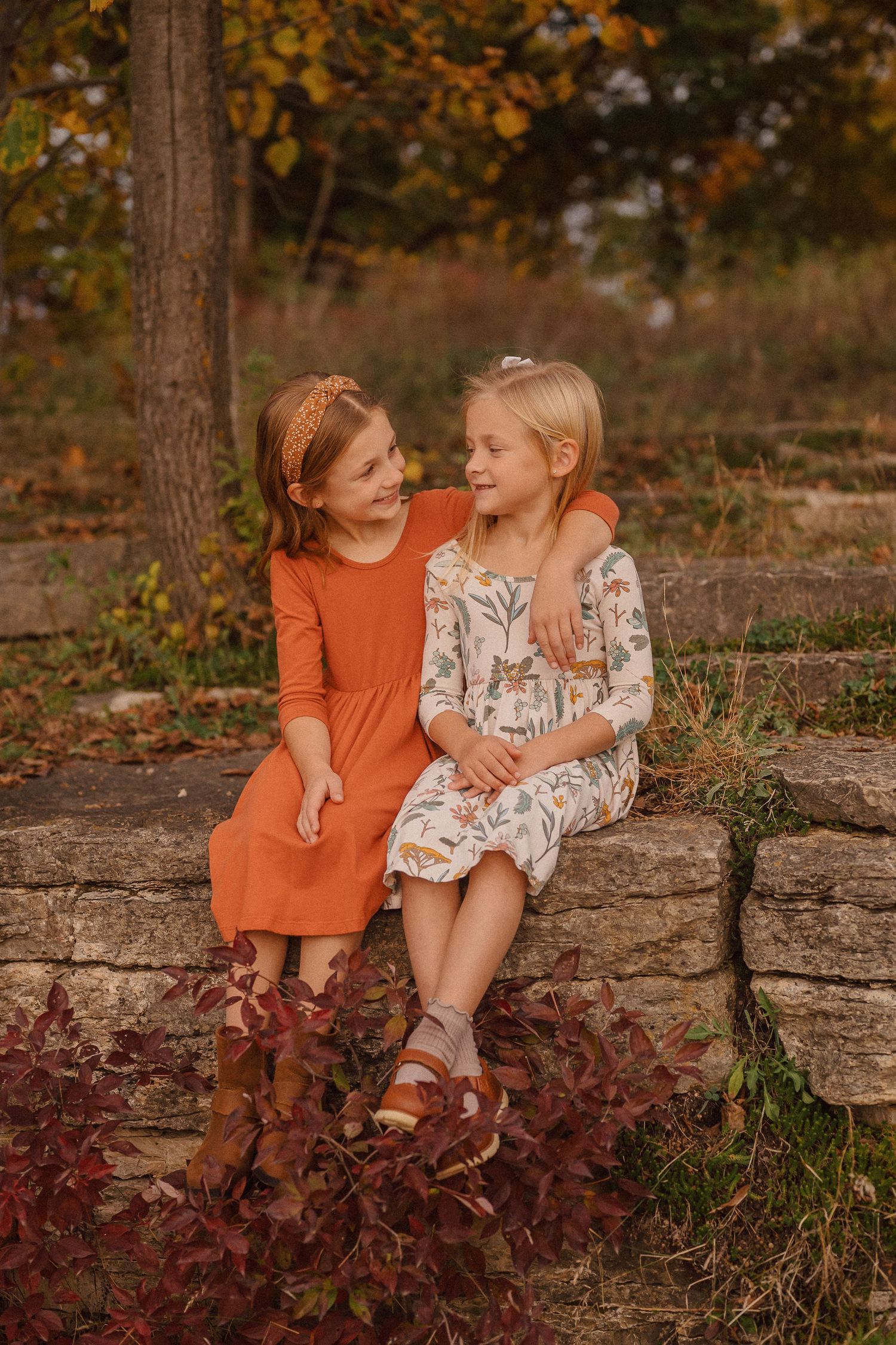 Two children in orange and floral dresses sit together on a stone wall surrounded by autumn colors.