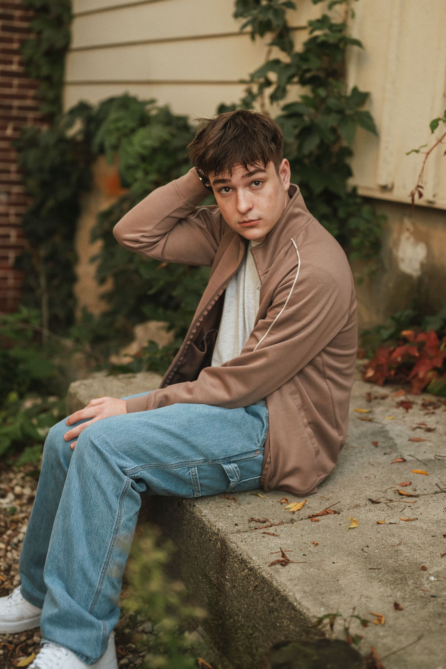 Person in brown jacket and jeans sitting on concrete ledge with brick wall and plants in background.