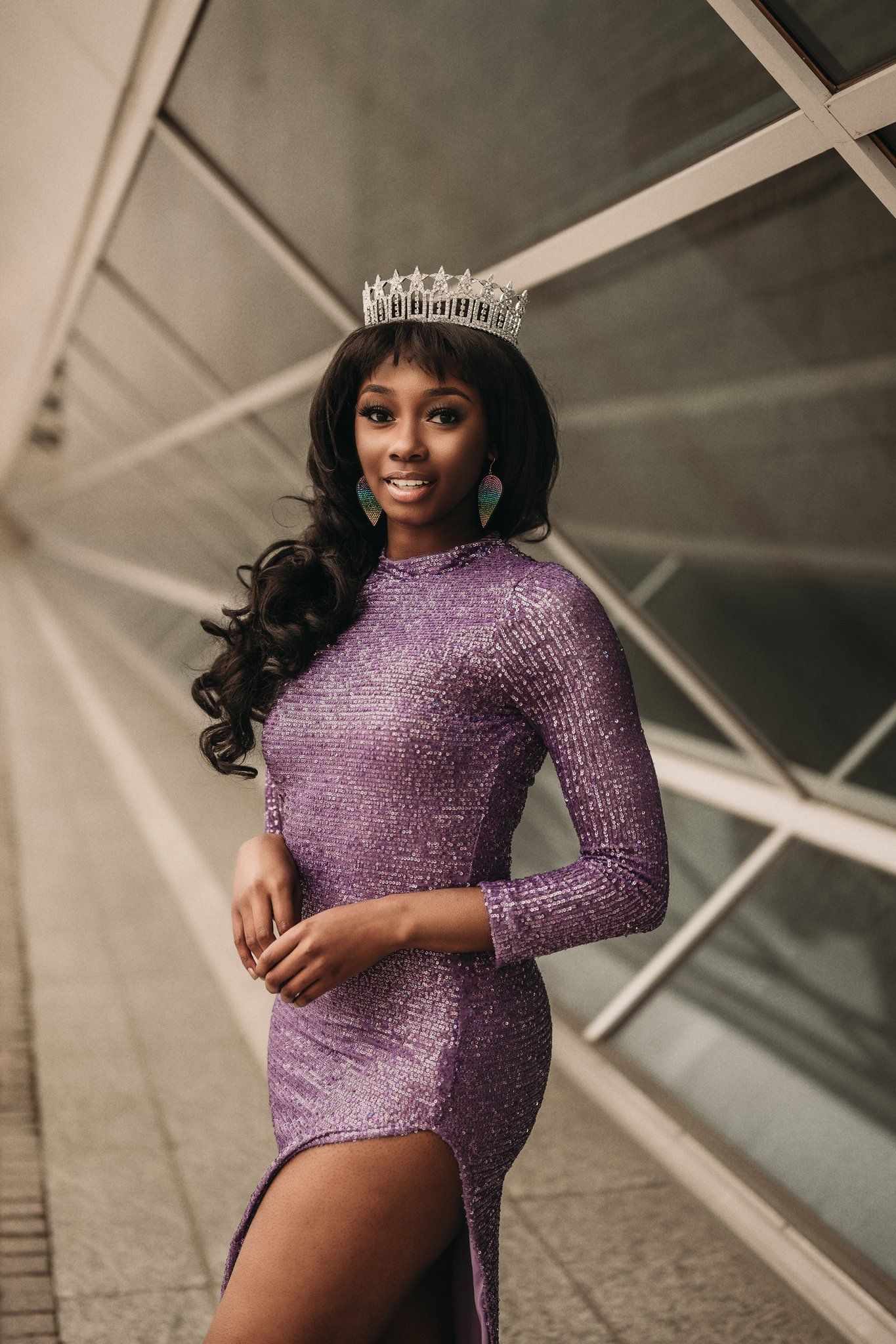 Beauty contestant in fitted purple gown poses against modern architectural backdrop.