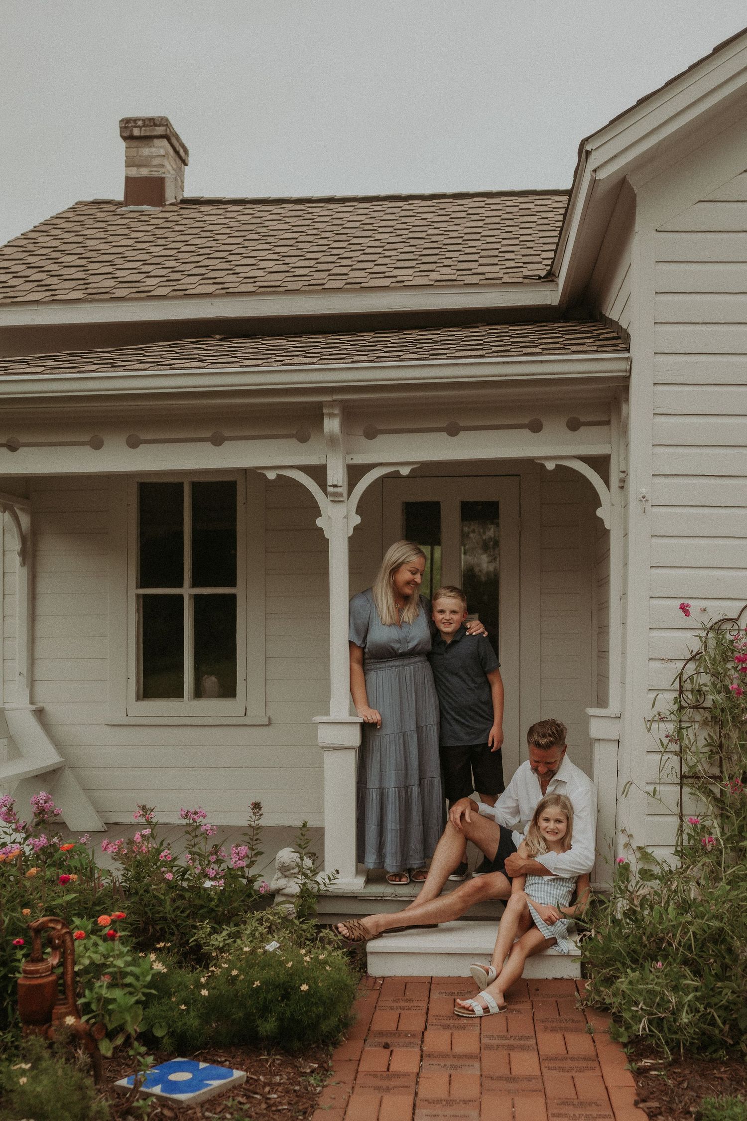 Family relaxes on front porch of classic white farmhouse with brick walkway.