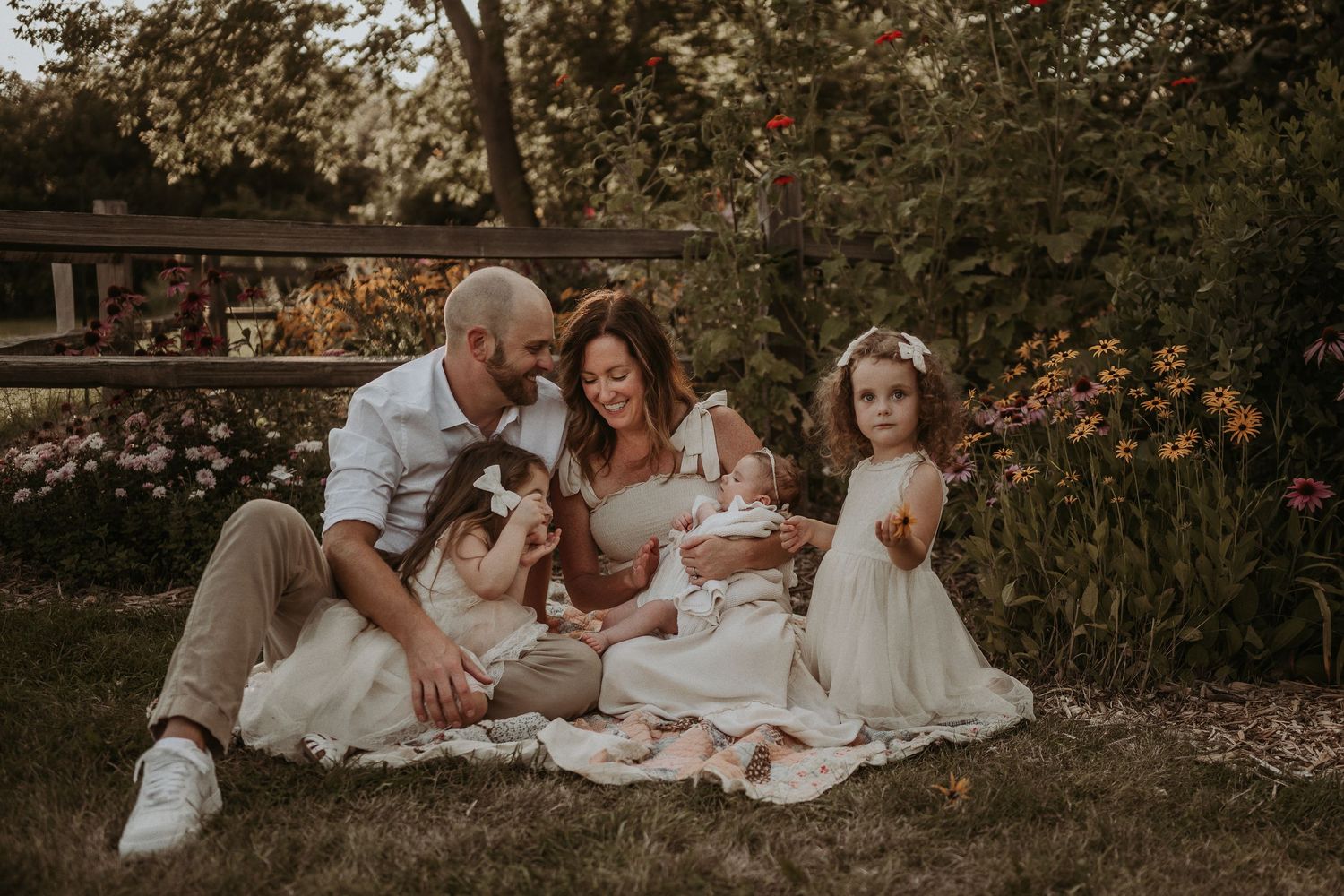 Family dressed in cream and white sits together on blanket in garden during sunset.