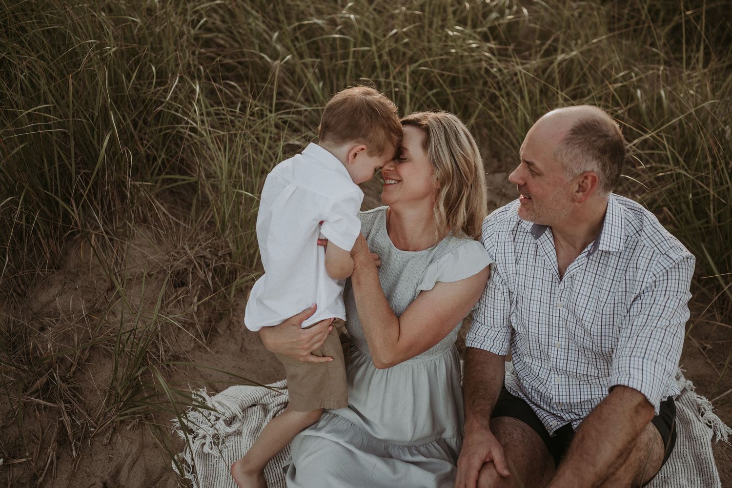 Tender family moment captured on sandy dunes with tall grass in background.