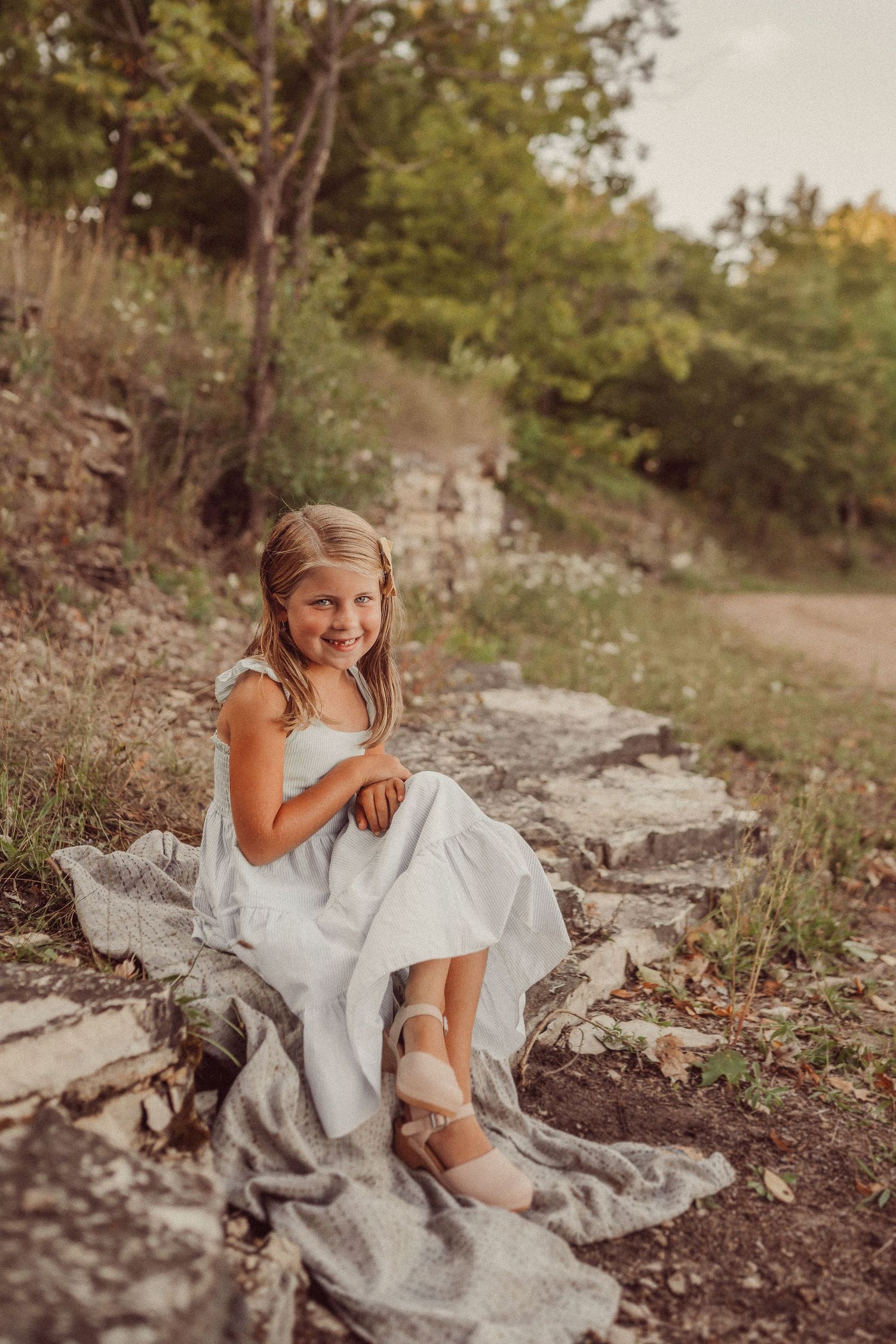 A child in a white dress sits on a blanket along a rocky hillside path.