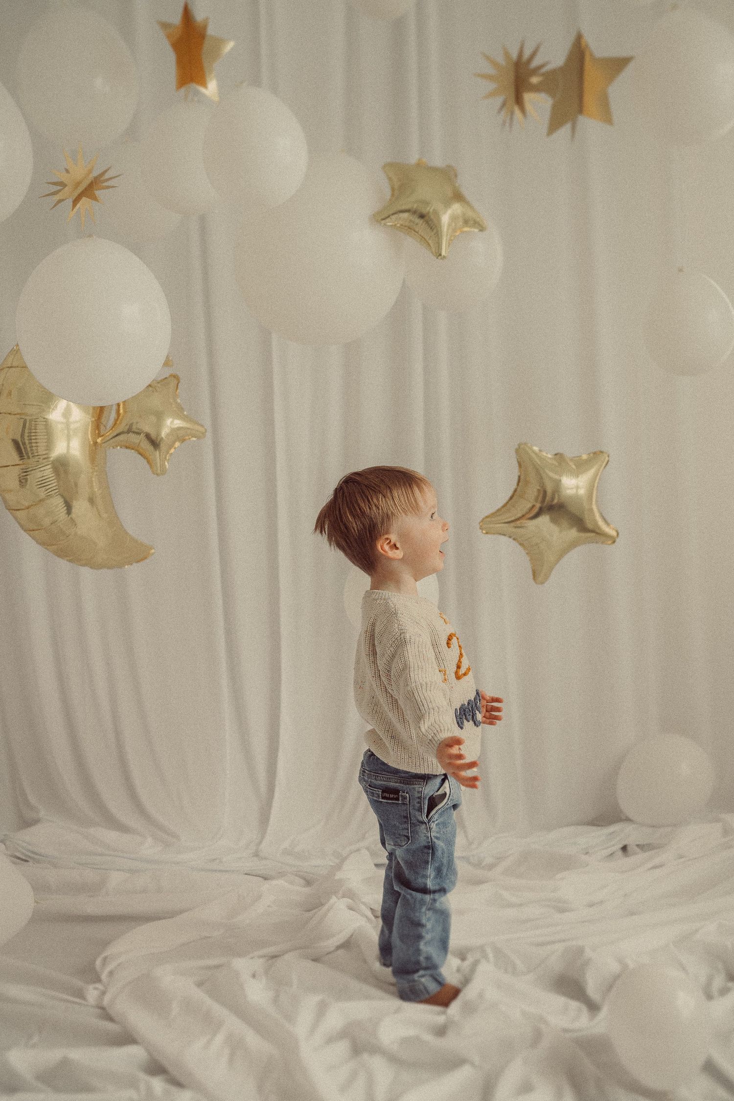 Child standing amidst white balloons and gold star decorations looking up at festive display.