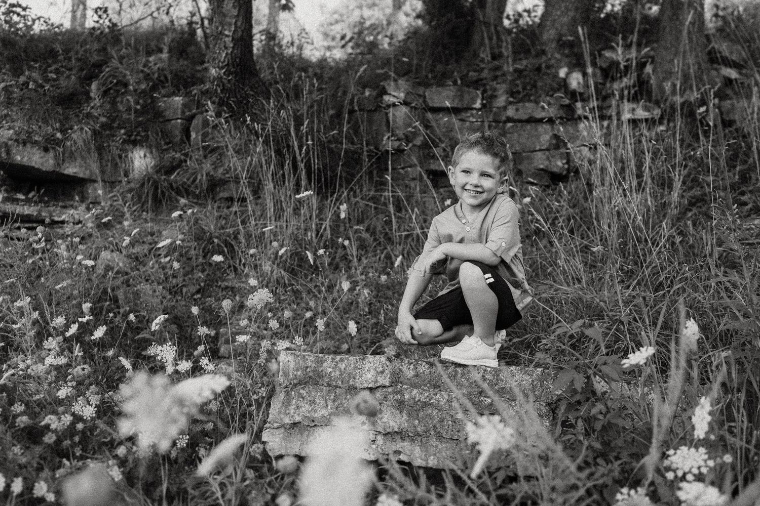 Black and white photo of child sitting on rocky ledge among wildflowers and tall grass.