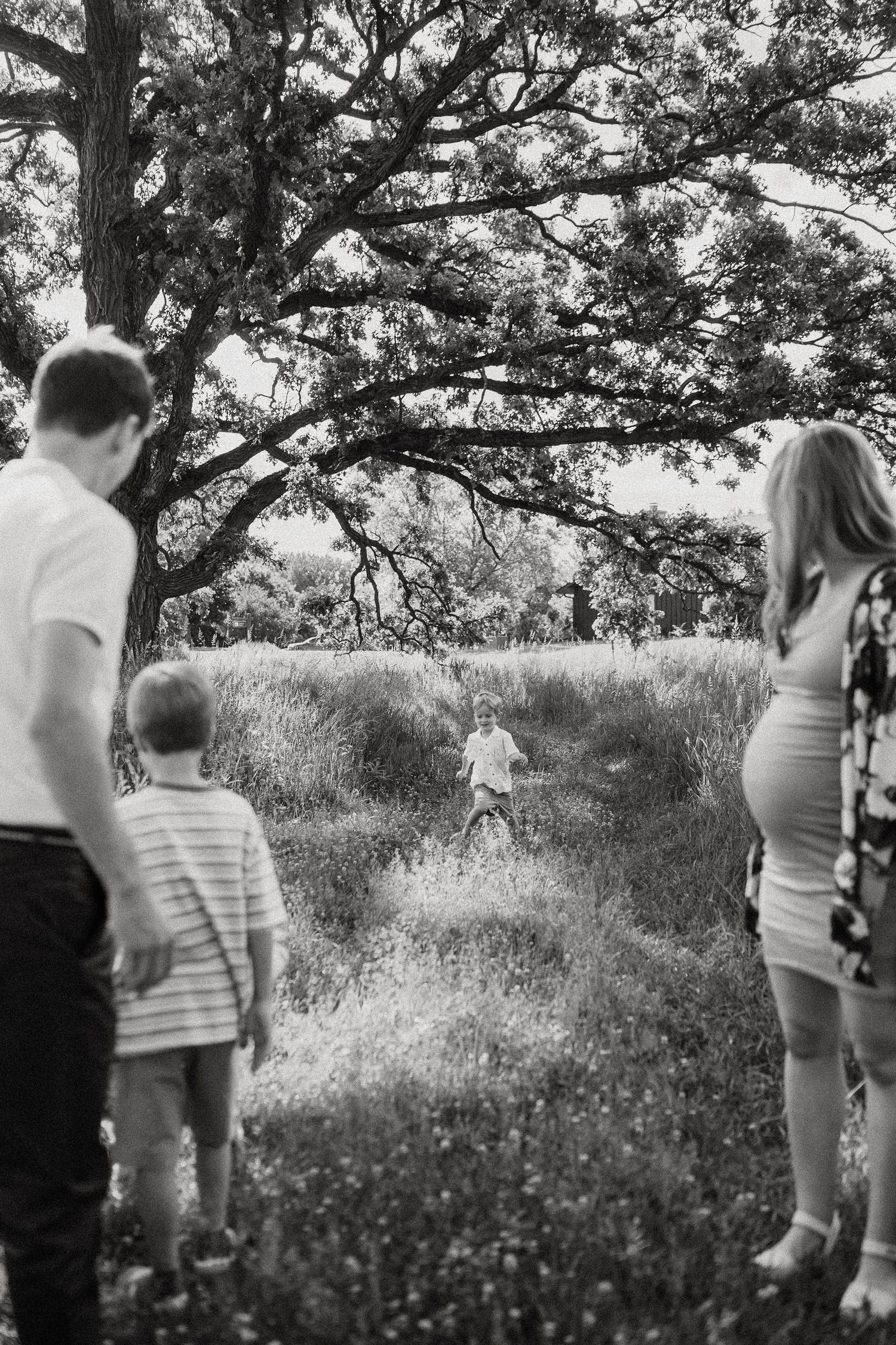 Black and white photo of a family watching two children play in a grassy field under a large tree.