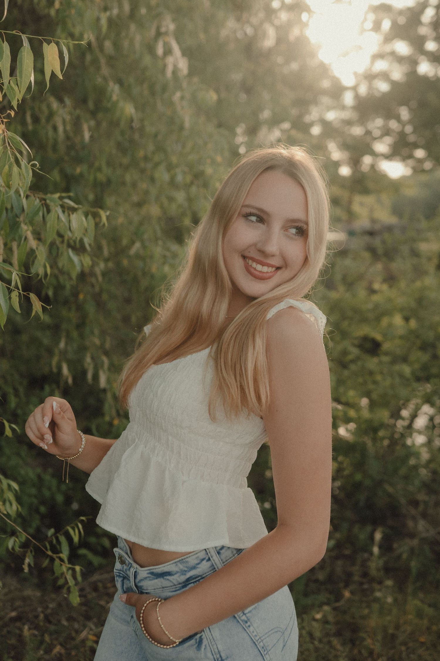 Smiling blonde woman in white crop top and jeans posing outdoors among lush green trees in golden sunlight.