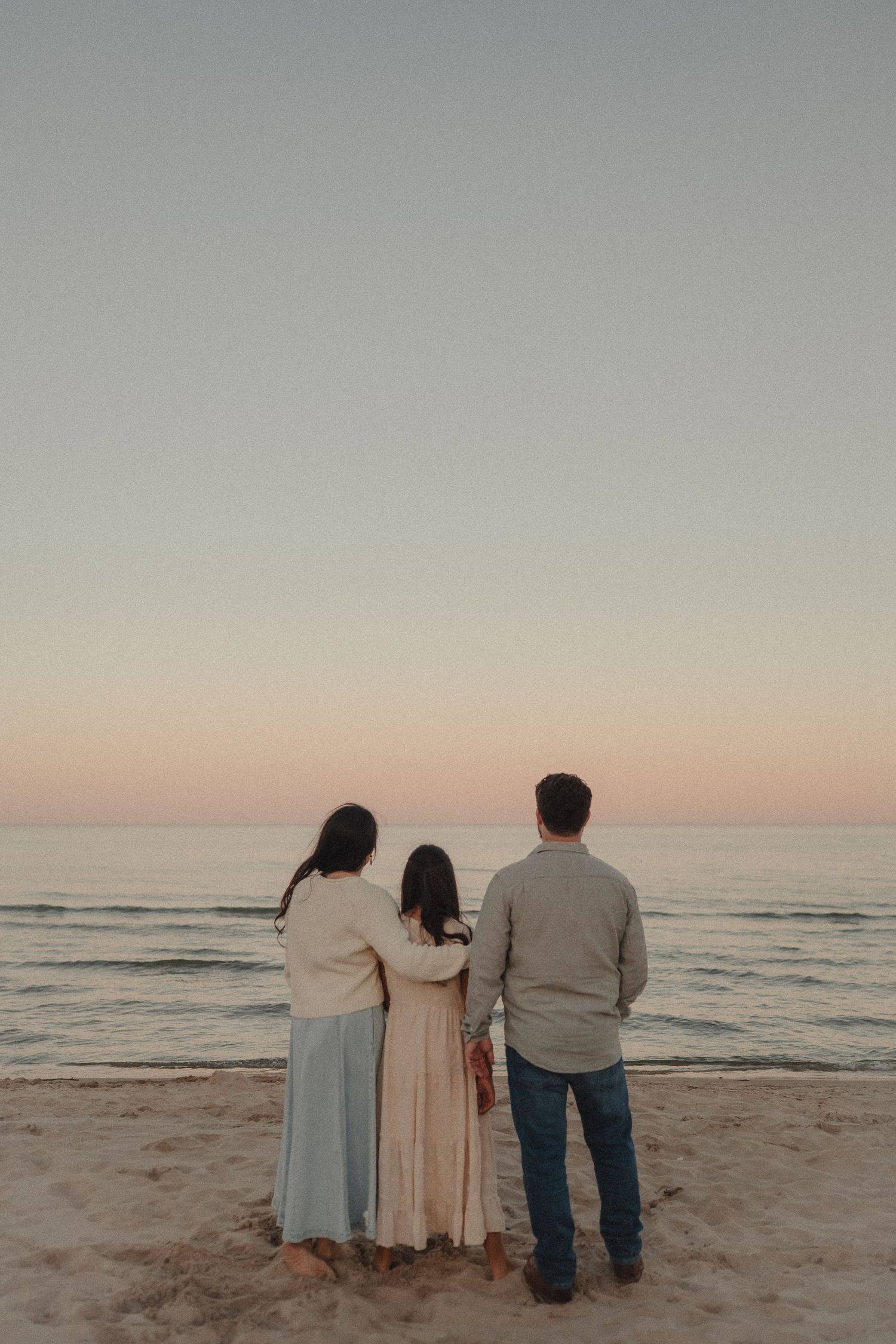 Family of three stands with backs to camera gazing at pastel sunset over ocean.