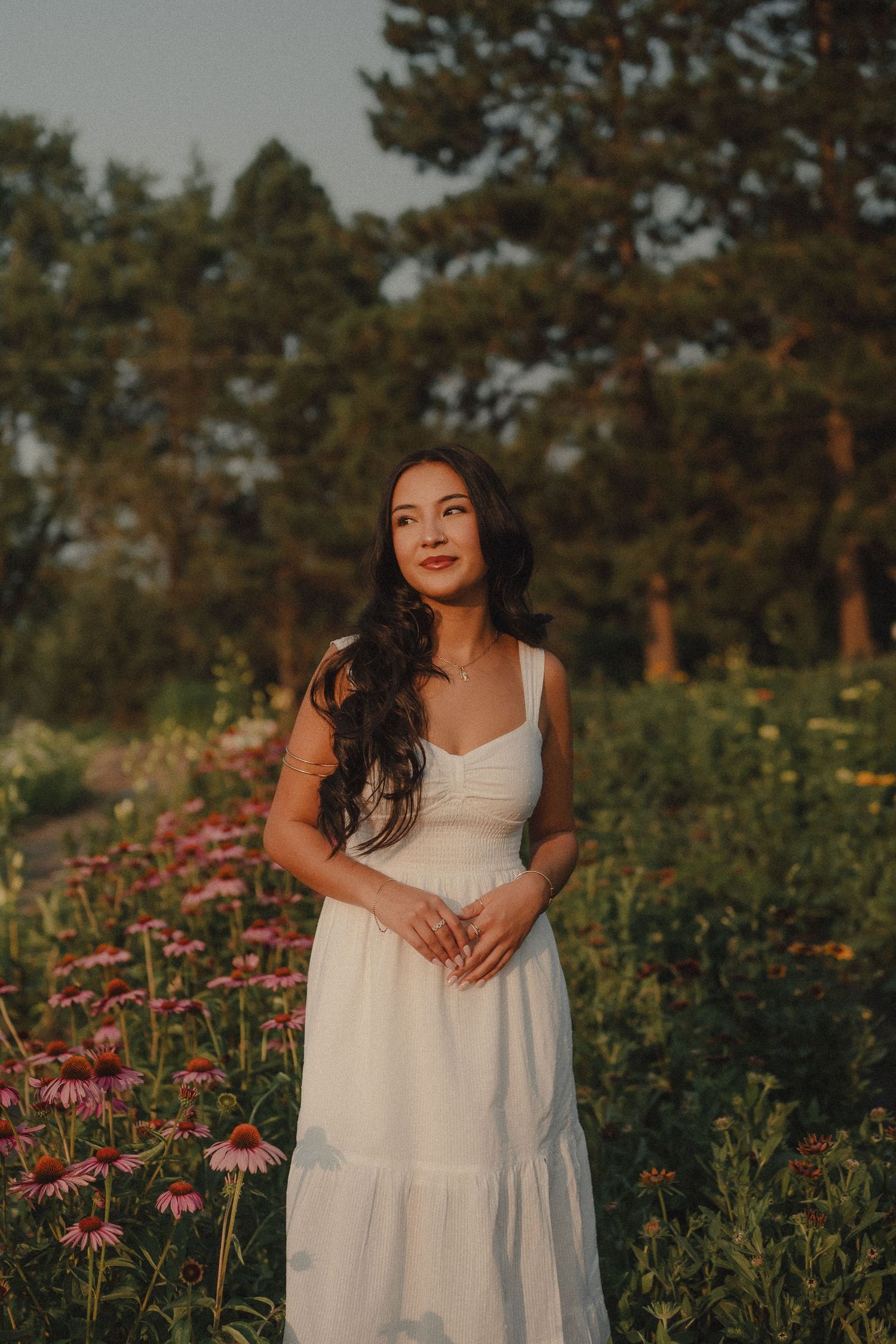 Woman in white dress standing in a flower garden at golden hour with lush green trees in the background.