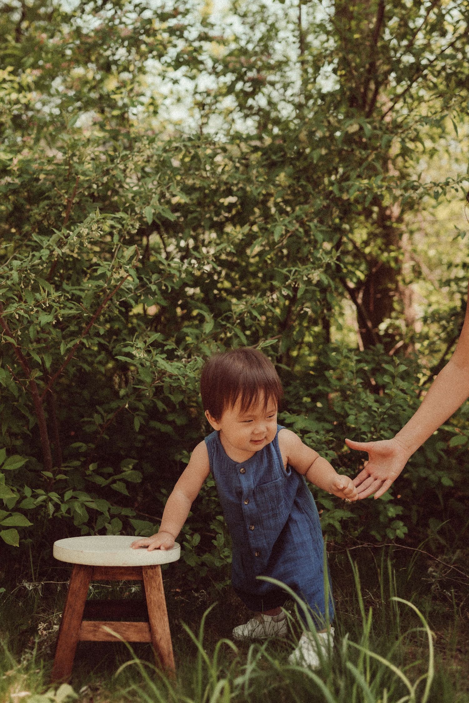 A toddler in denim overalls reaches for support while standing by a wooden stool in a garden setting.