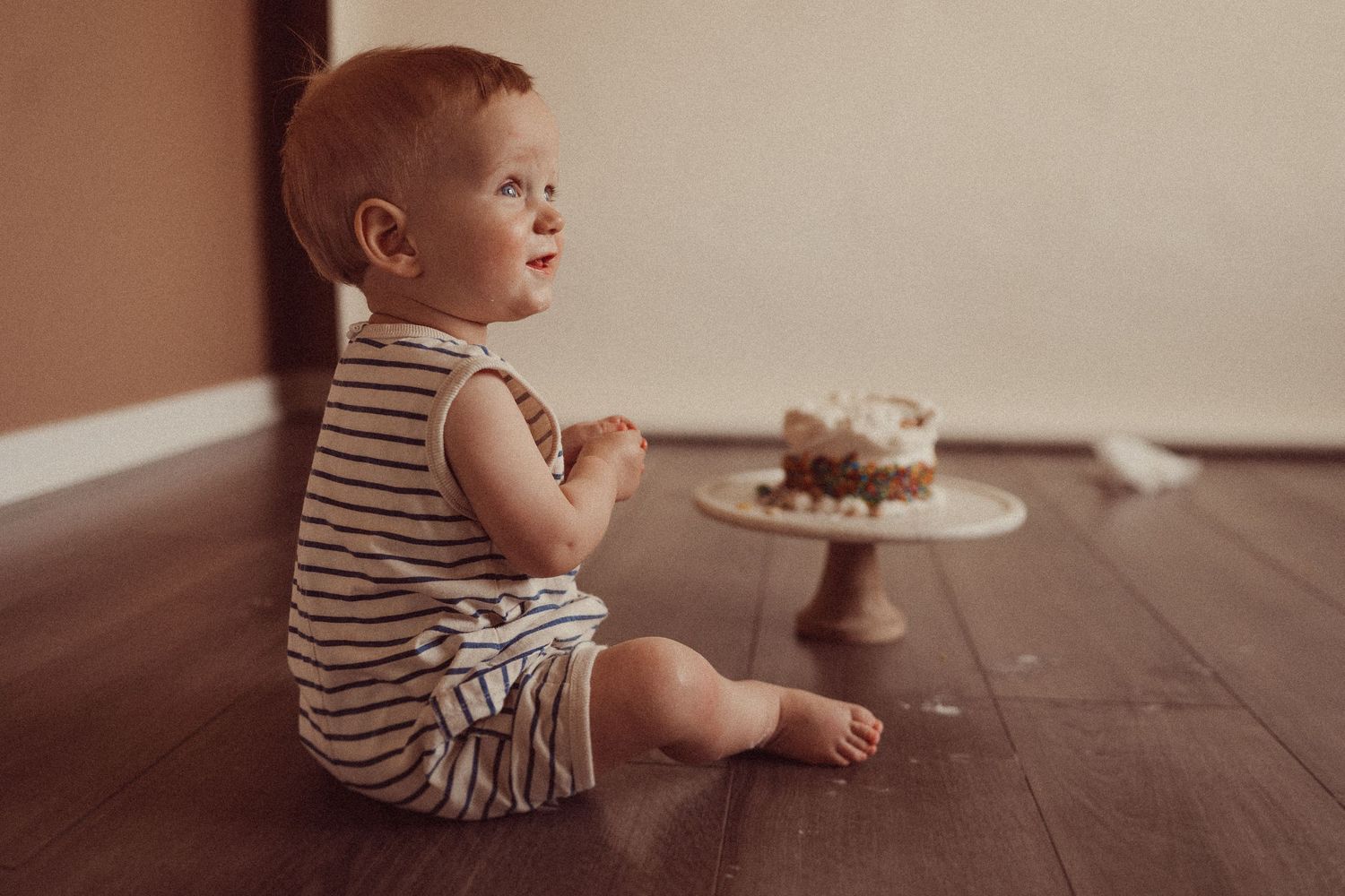 Sweet vintage-toned photo of toddler in striped outfit sitting near cake on wooden floor.