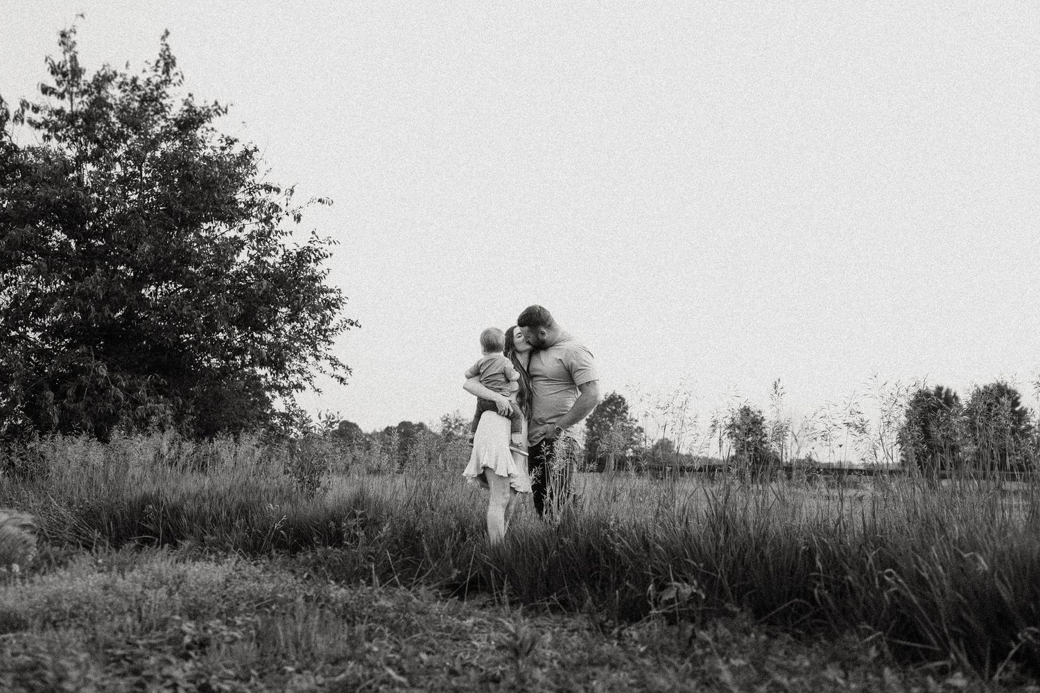 Black and white portrait of a tender family moment in a serene outdoor setting with natural vegetation.