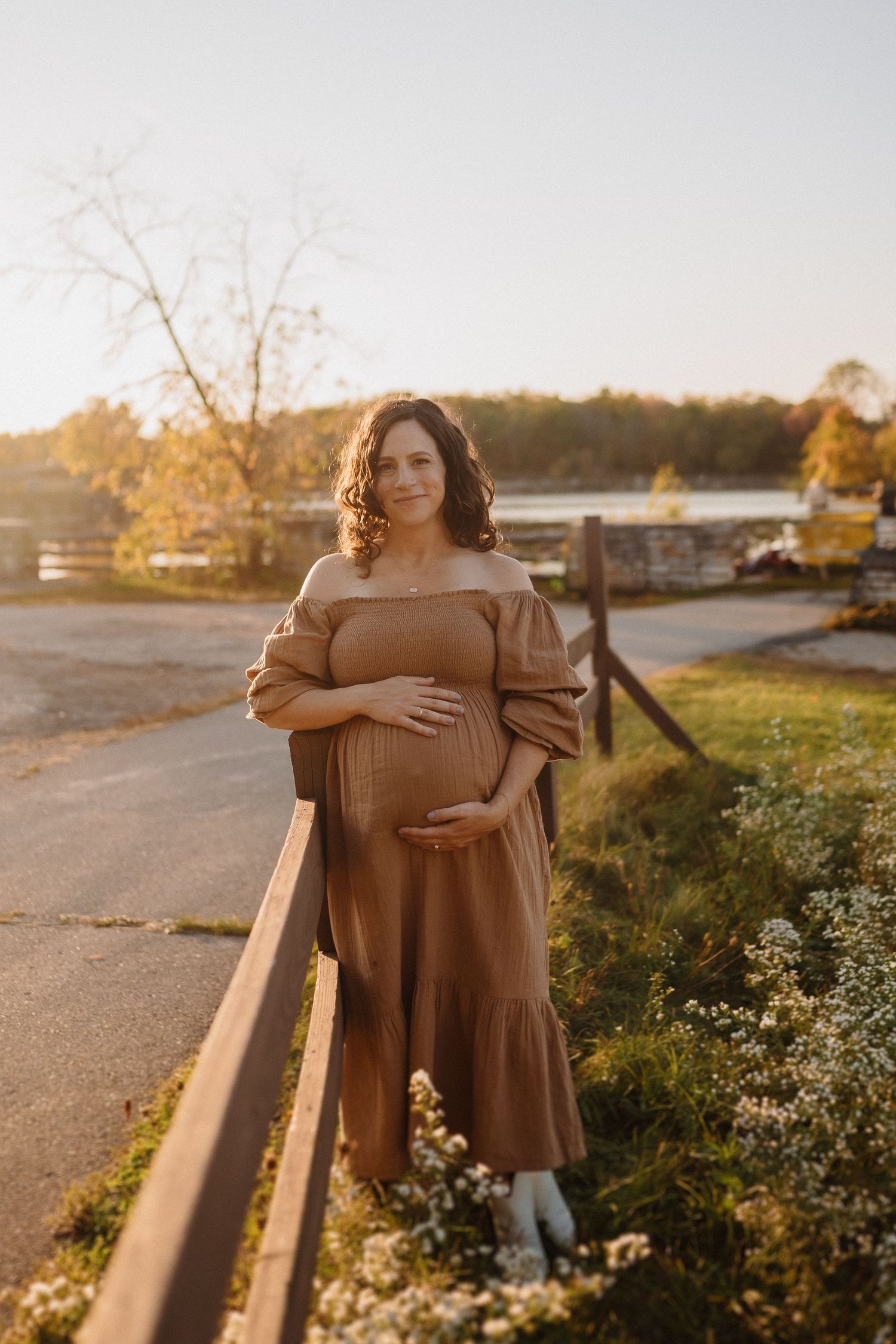 Maternity photo of woman in brown dress standing by wooden fence at sunset.