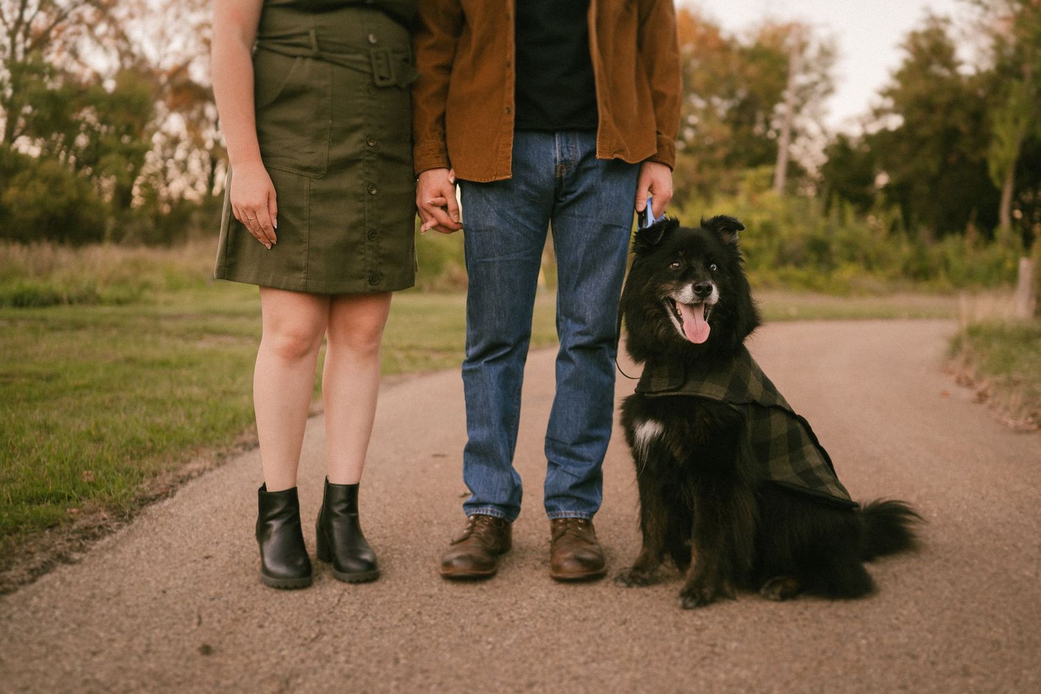 A black dog sits beside its owners' legs on a paved walking path.