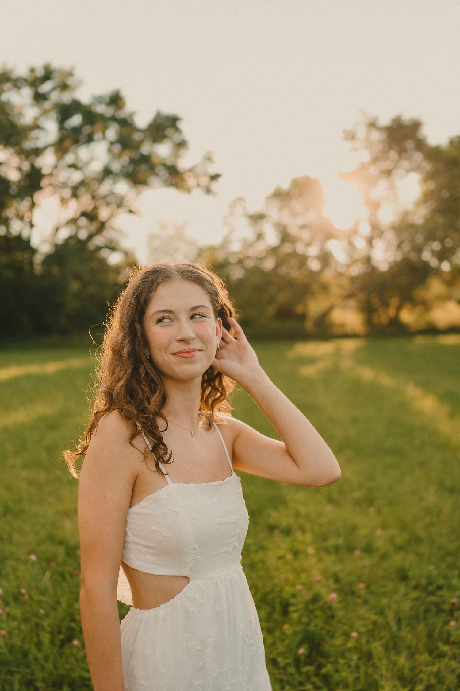 A person in a white cut-out dress poses in a field during golden hour with a dreamy sunset backdrop.