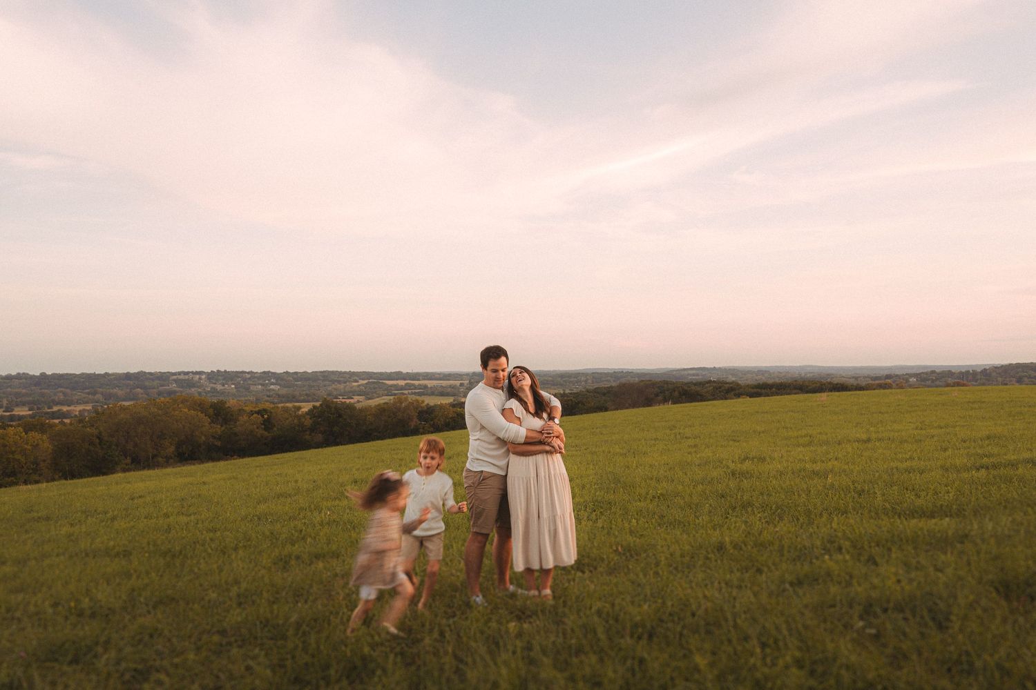 A family embraces on a grassy hilltop during golden hour with their dog running in the background.